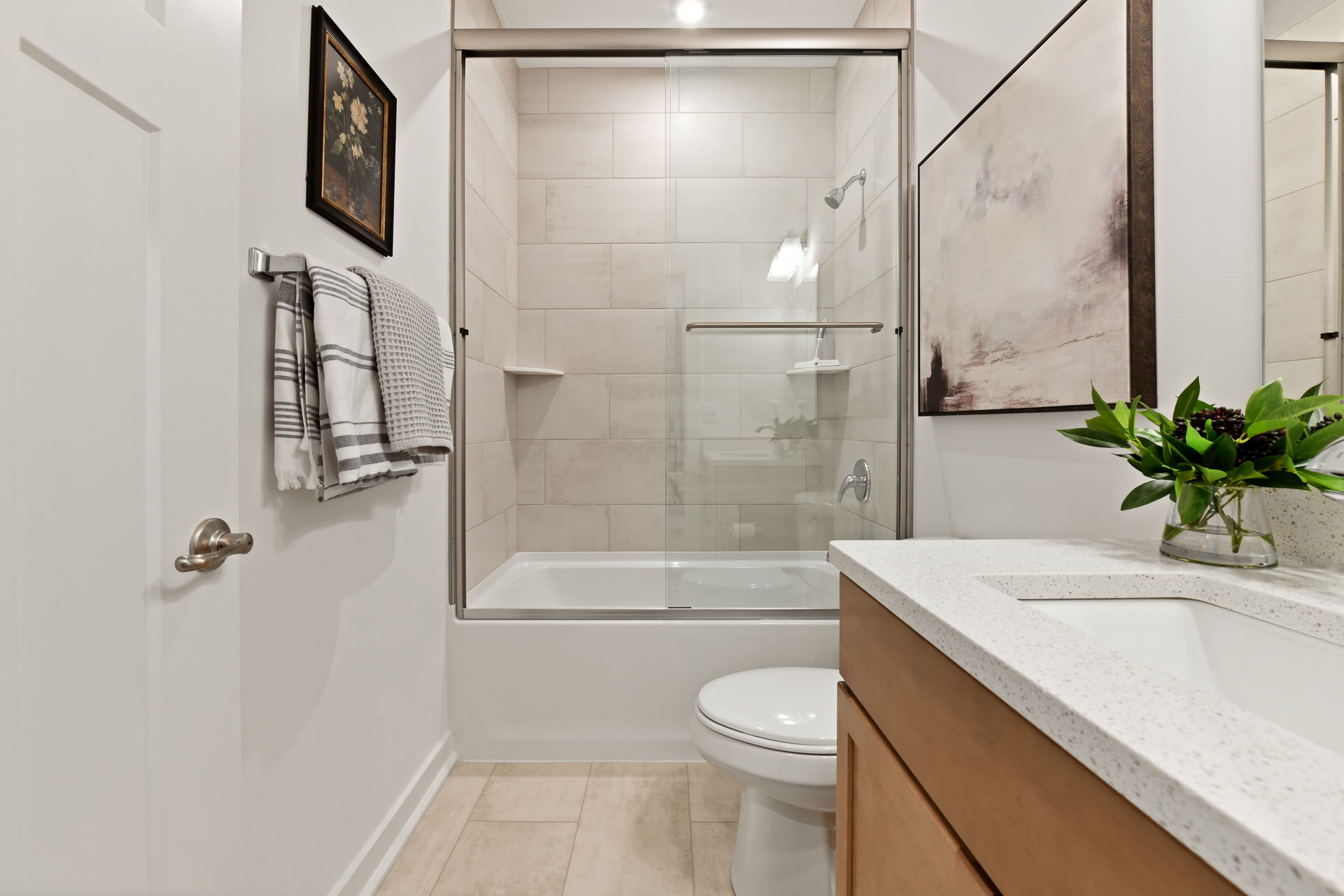 Modern bathroom with white tiles, a glass shower door, wooden vanity, and decorative wall art.