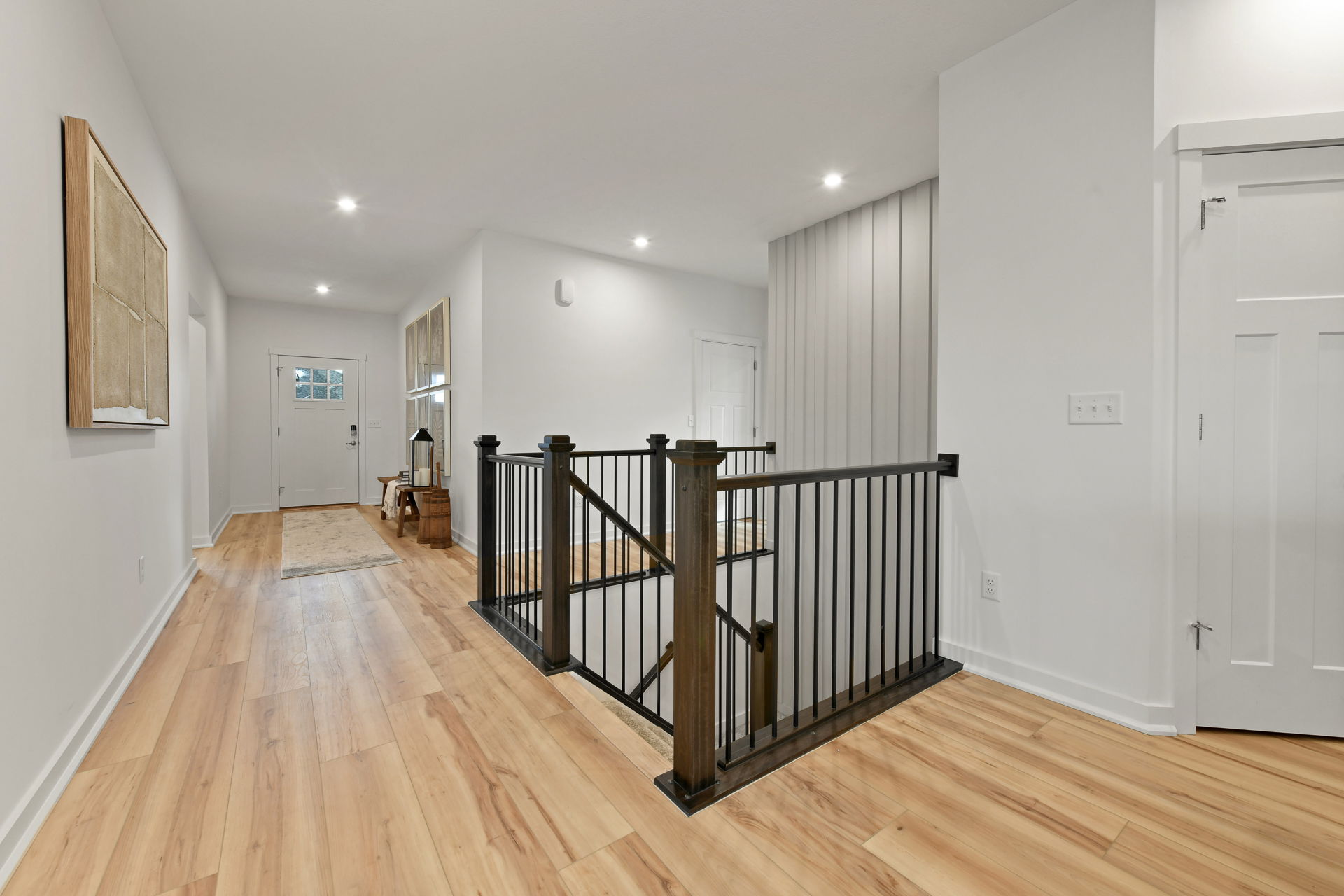 Spacious modern hallway with light wood flooring, contemporary railing, and white walls leading to a closed front door.