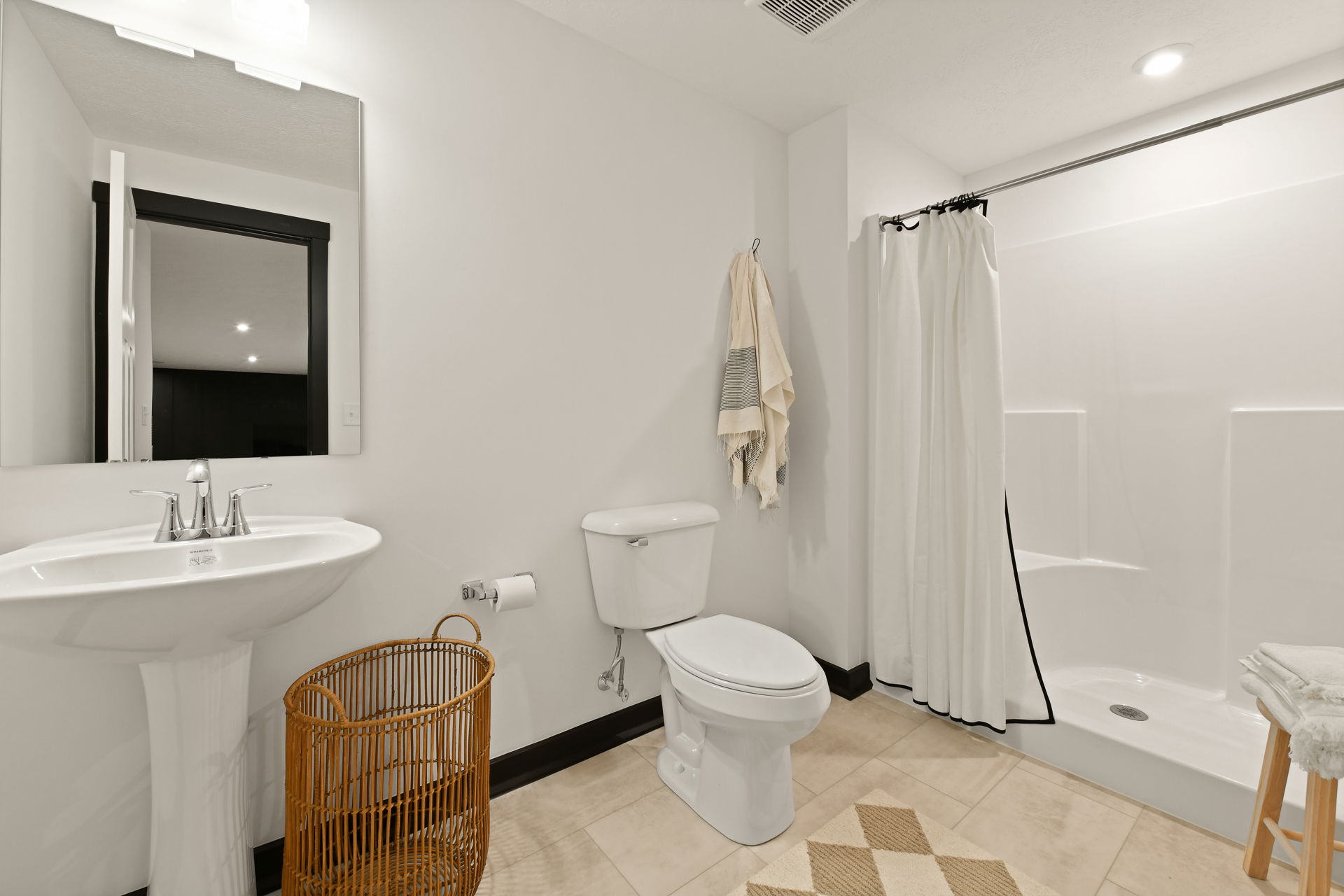 Modern bathroom with white fixtures, pedestal sink, wicker basket, wall mirror, and a shower with a curtain.