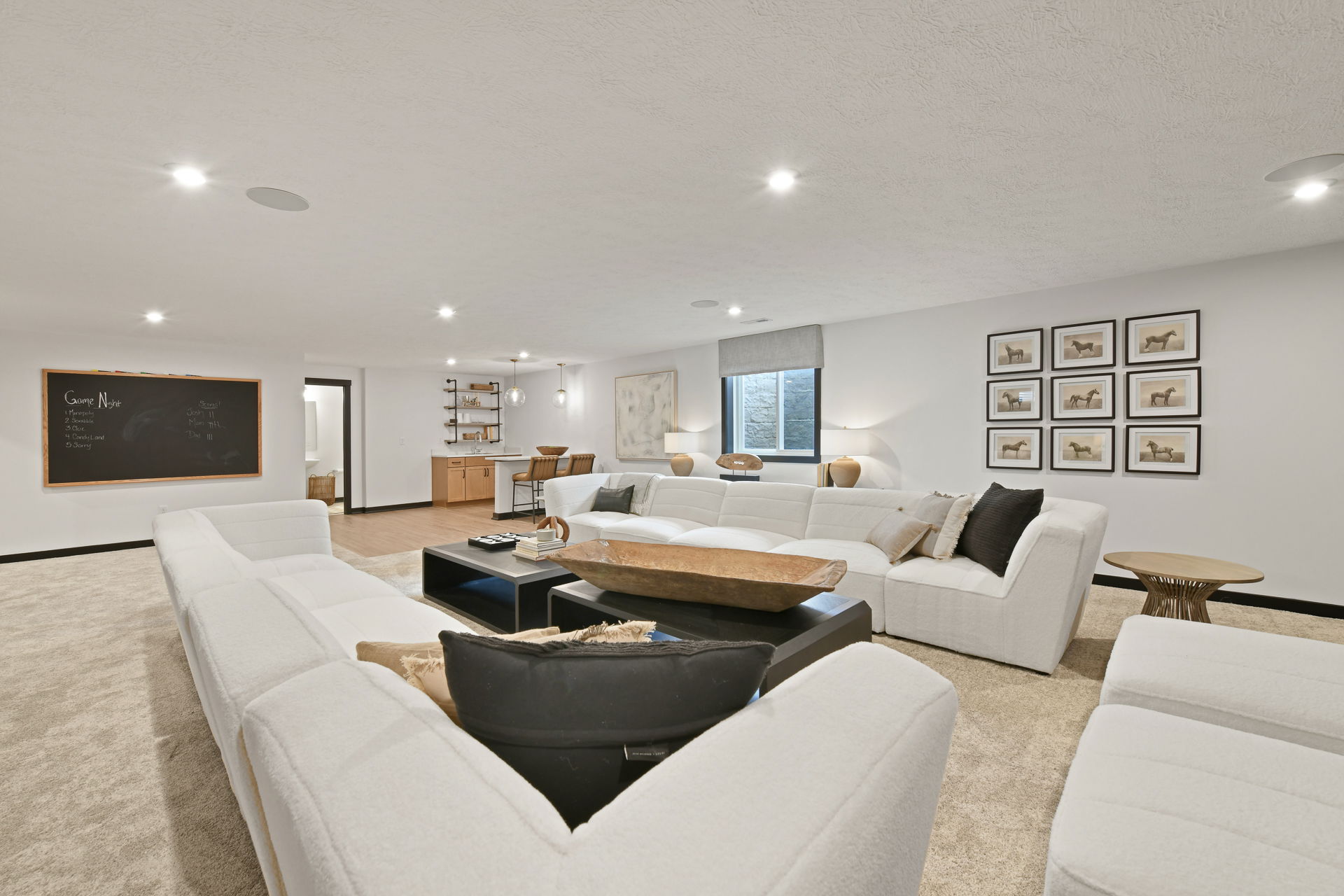 Spacious modern living room with white sectional sofas, a black coffee table, wall art, and a game night chalkboard, featuring cozy lighting and a bar area.