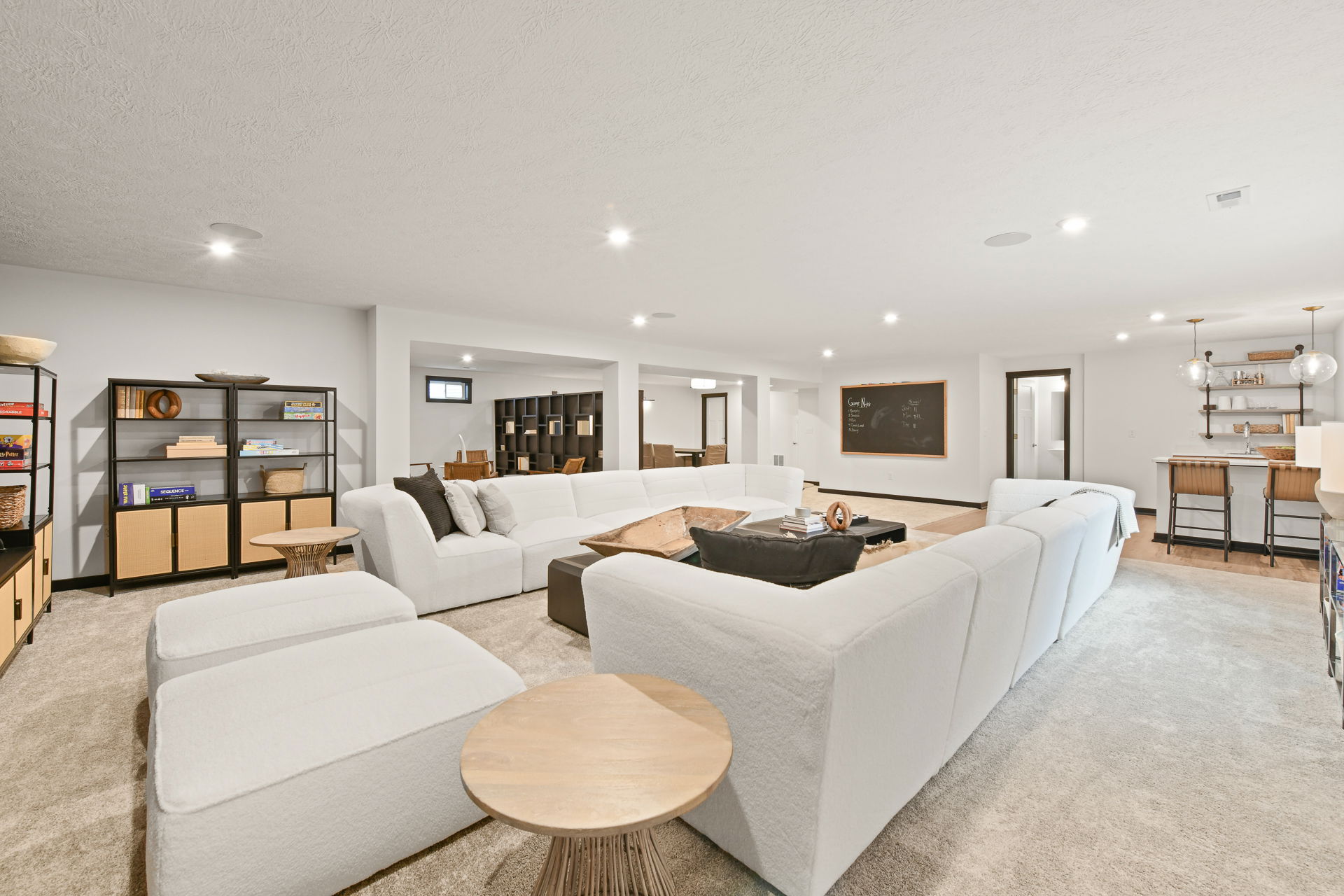 Spacious modern basement living area with white sectional sofa, cozy carpet, bookshelves, and a small bar area featuring barstools and pendant lighting.