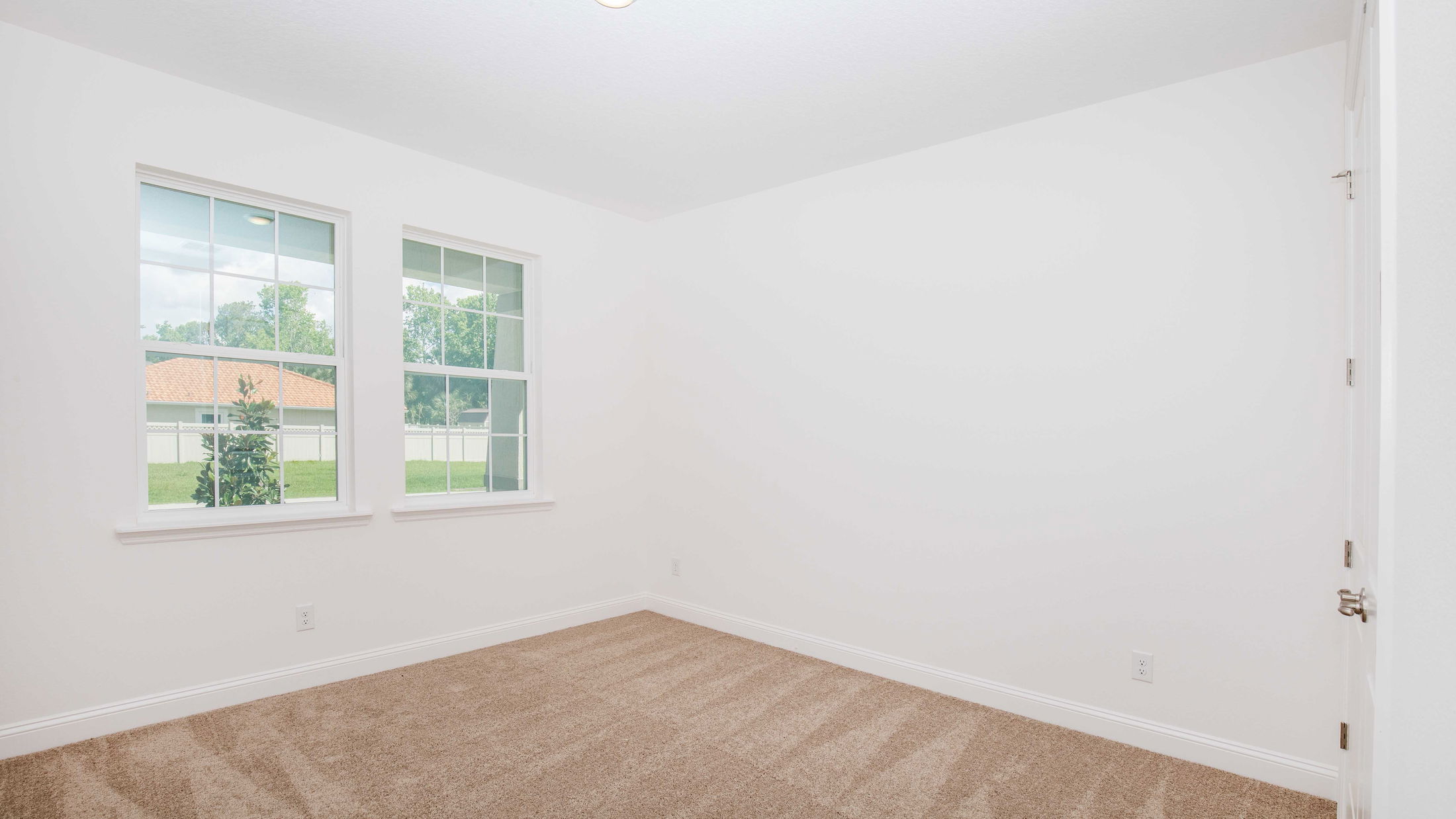 Empty room with beige carpet, white walls, and two large windows overlooking a green backyard.