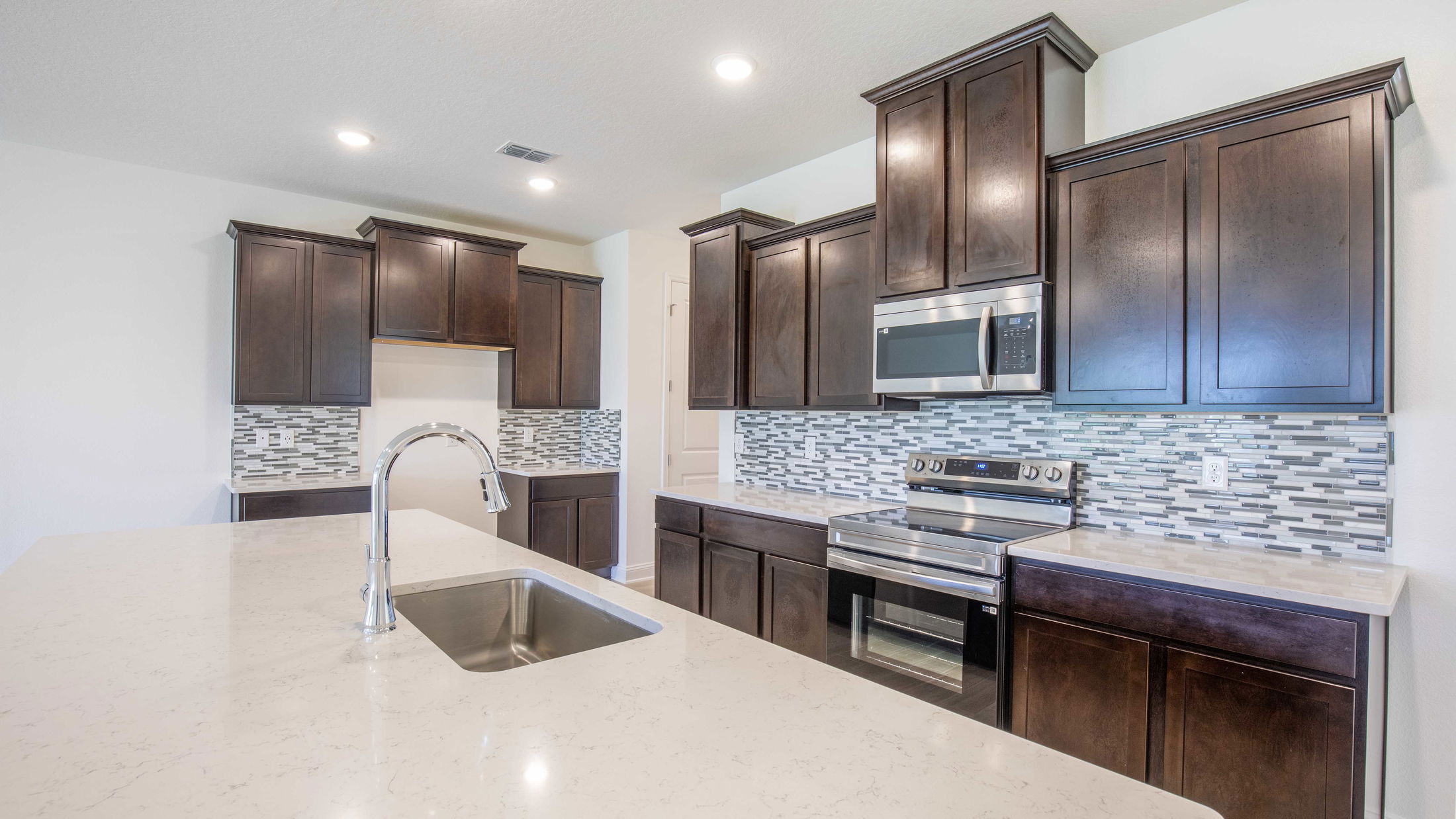Modern kitchen with dark wood cabinets, stainless steel appliances, a light quartz countertop, and a gray mosaic tile backsplash.