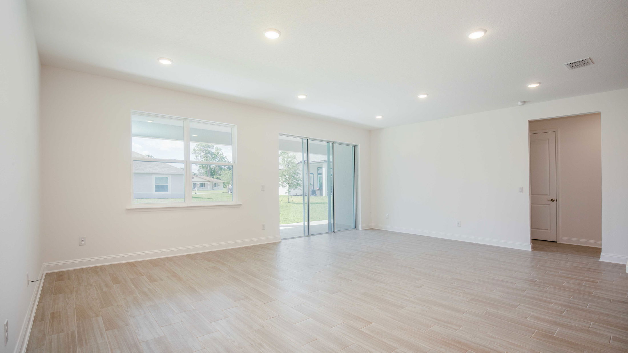 Spacious and bright empty living room with beige wooden flooring, recessed lighting, and large windows overlooking a green lawn.