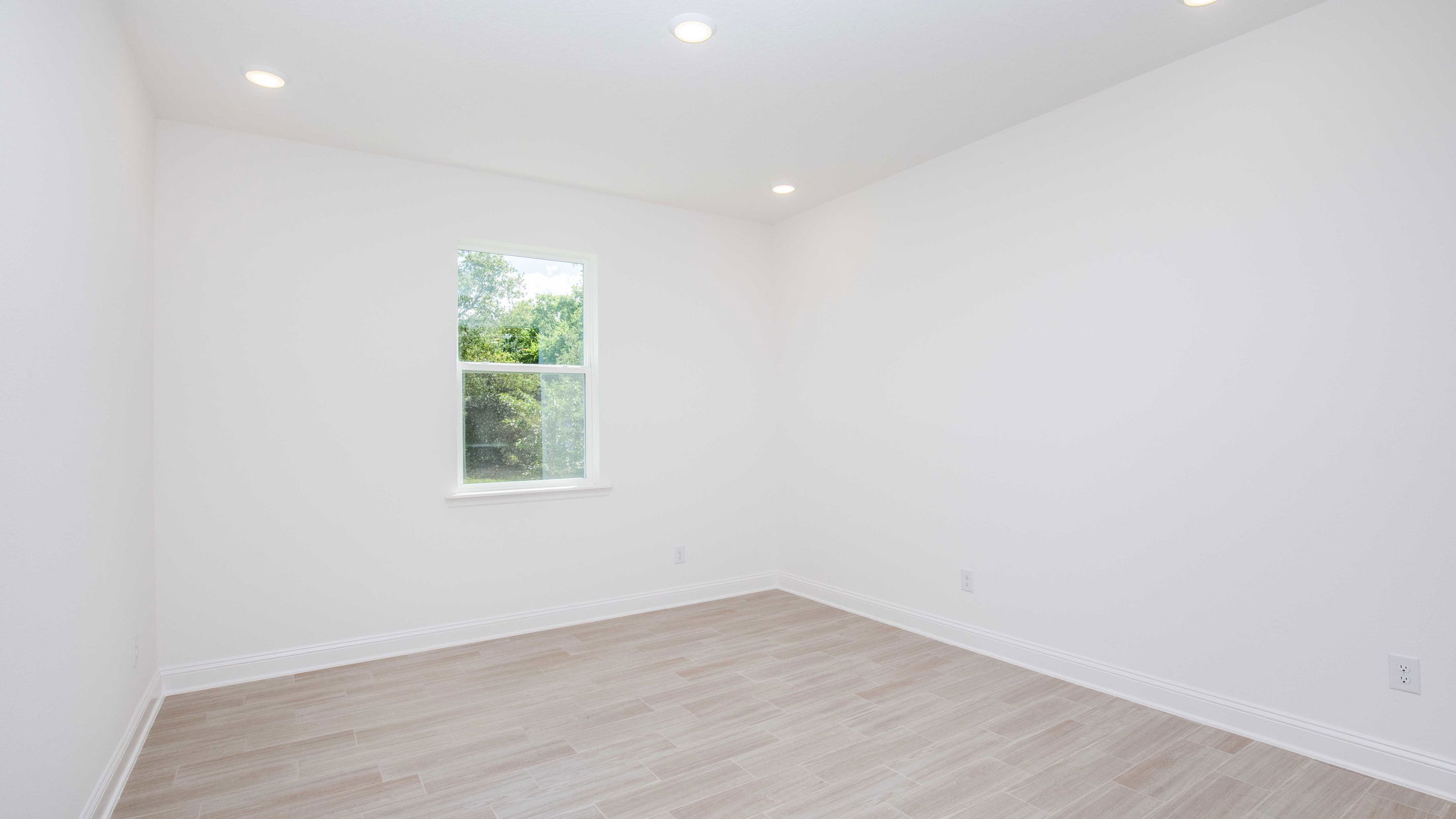Bright empty room with white walls, hardwood flooring, recessed ceiling lights, and a window overlooking greenery.