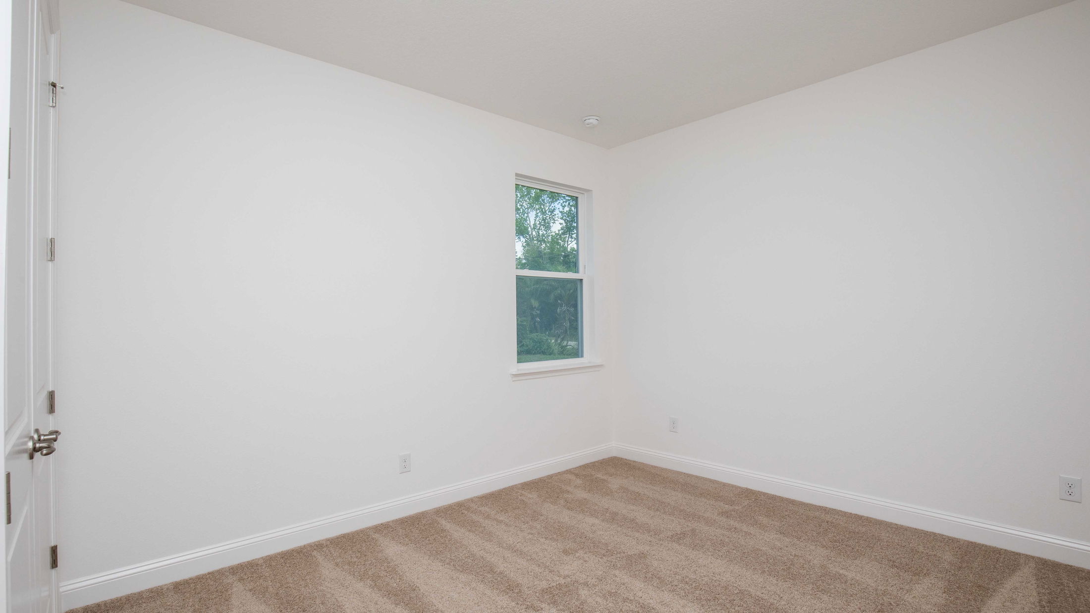 A small, empty room with white walls, a single window, and beige carpet flooring.