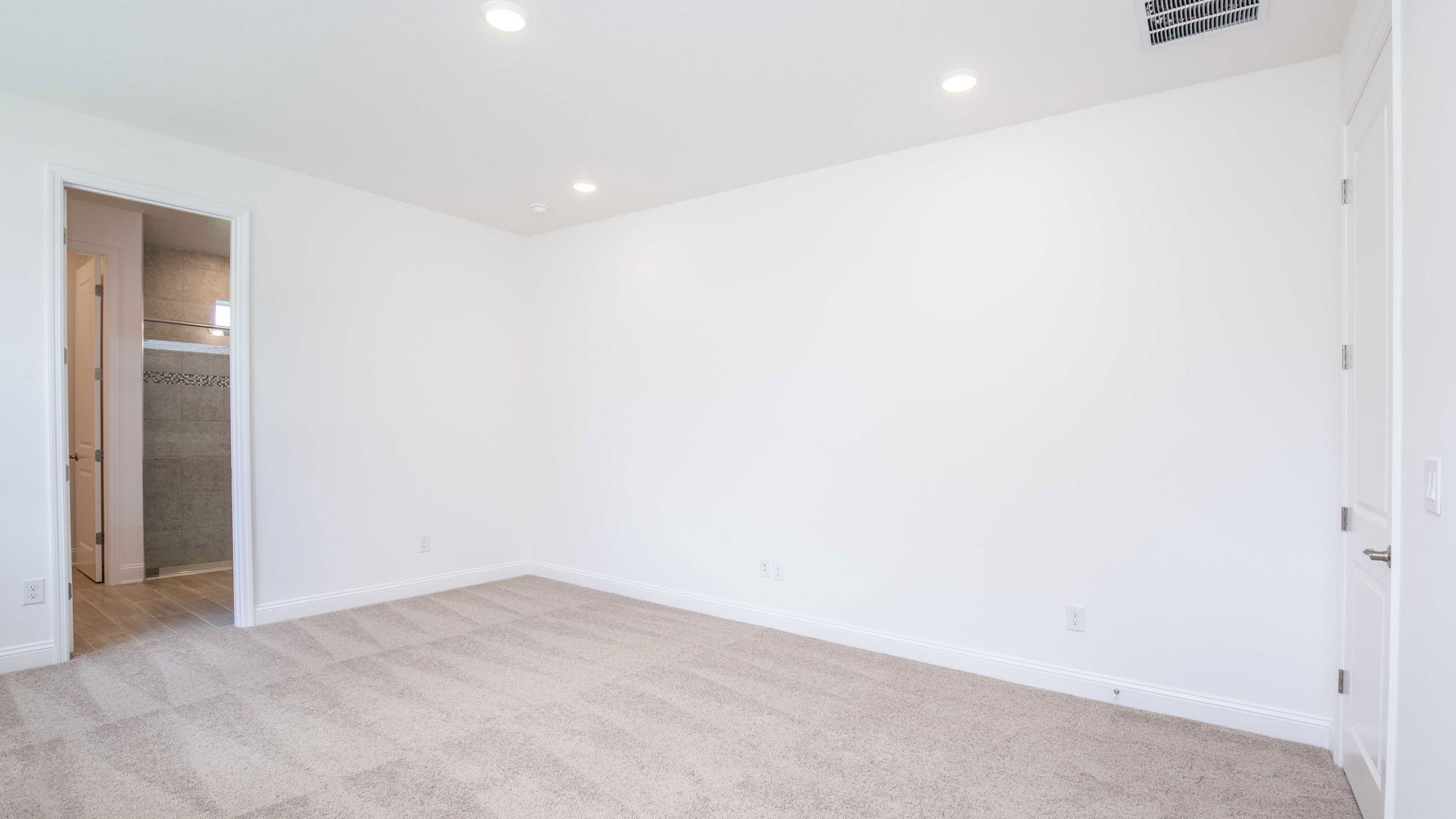 Empty white room with beige carpet flooring and an open door leading to a tiled bathroom.
