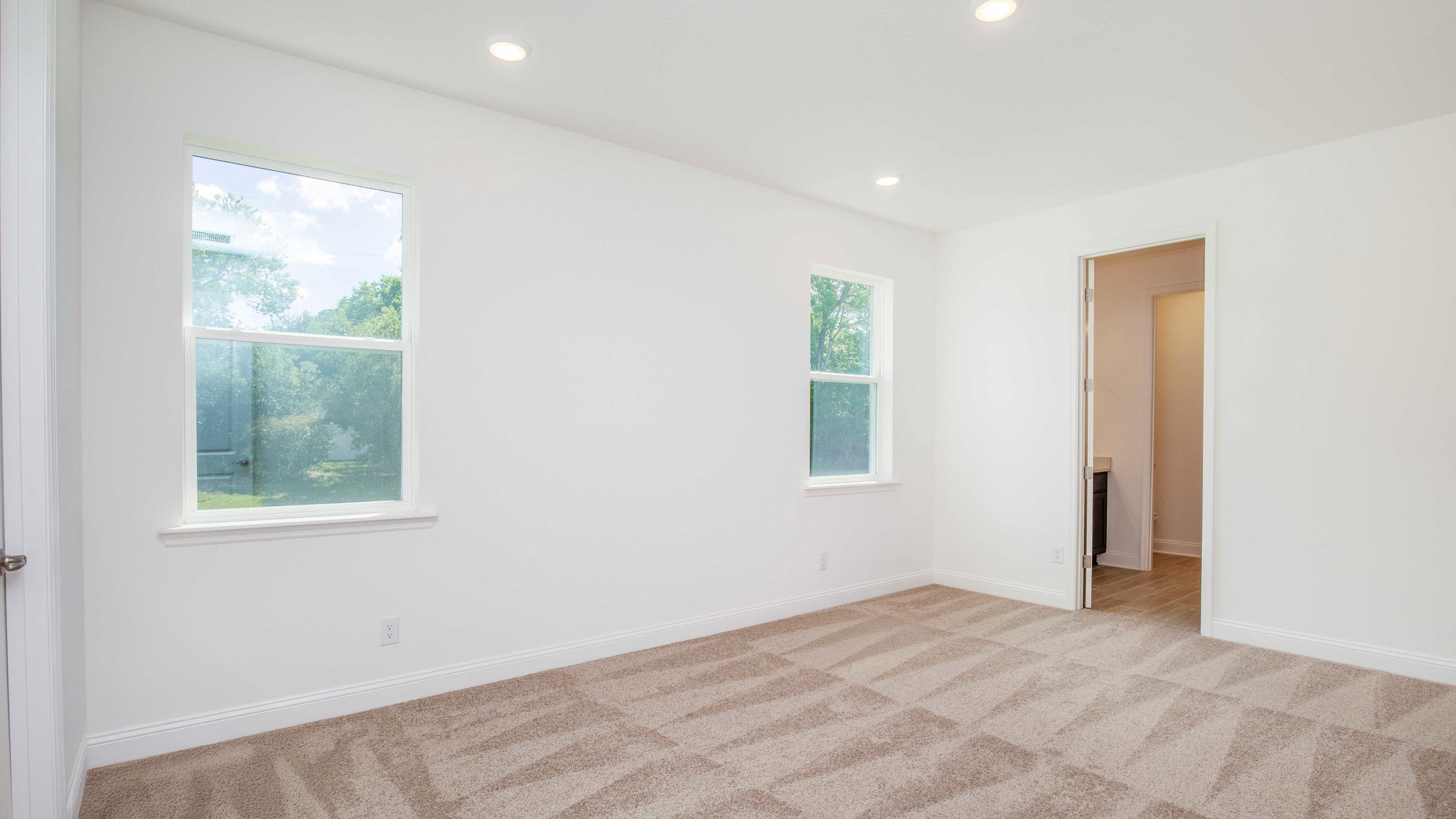 Bright empty room with white walls, beige carpet, two windows, and an open door leading to a hallway in a modern home.
