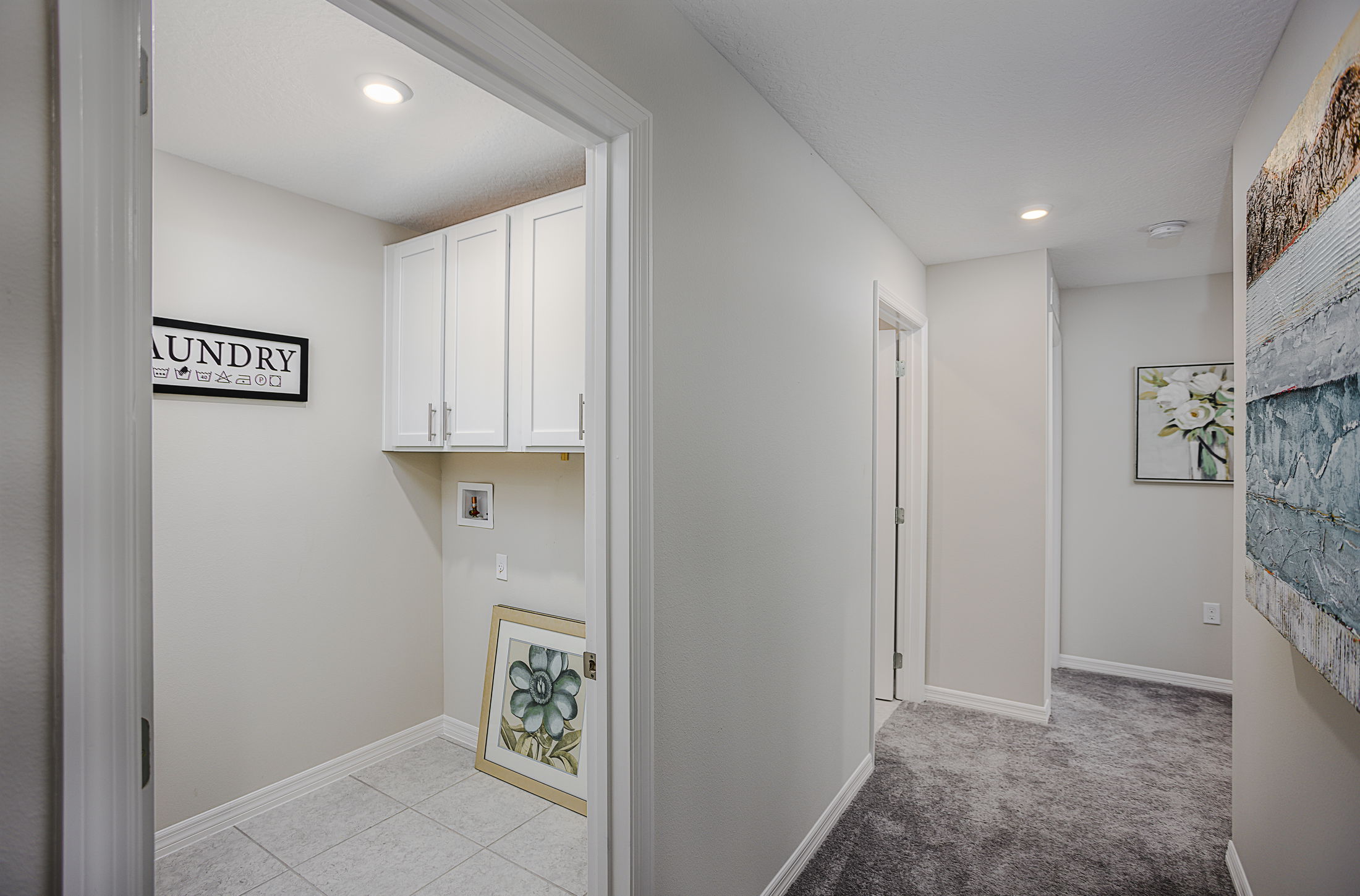 Hallway leading to a compact laundry room with white cabinets and decorative artwork.
