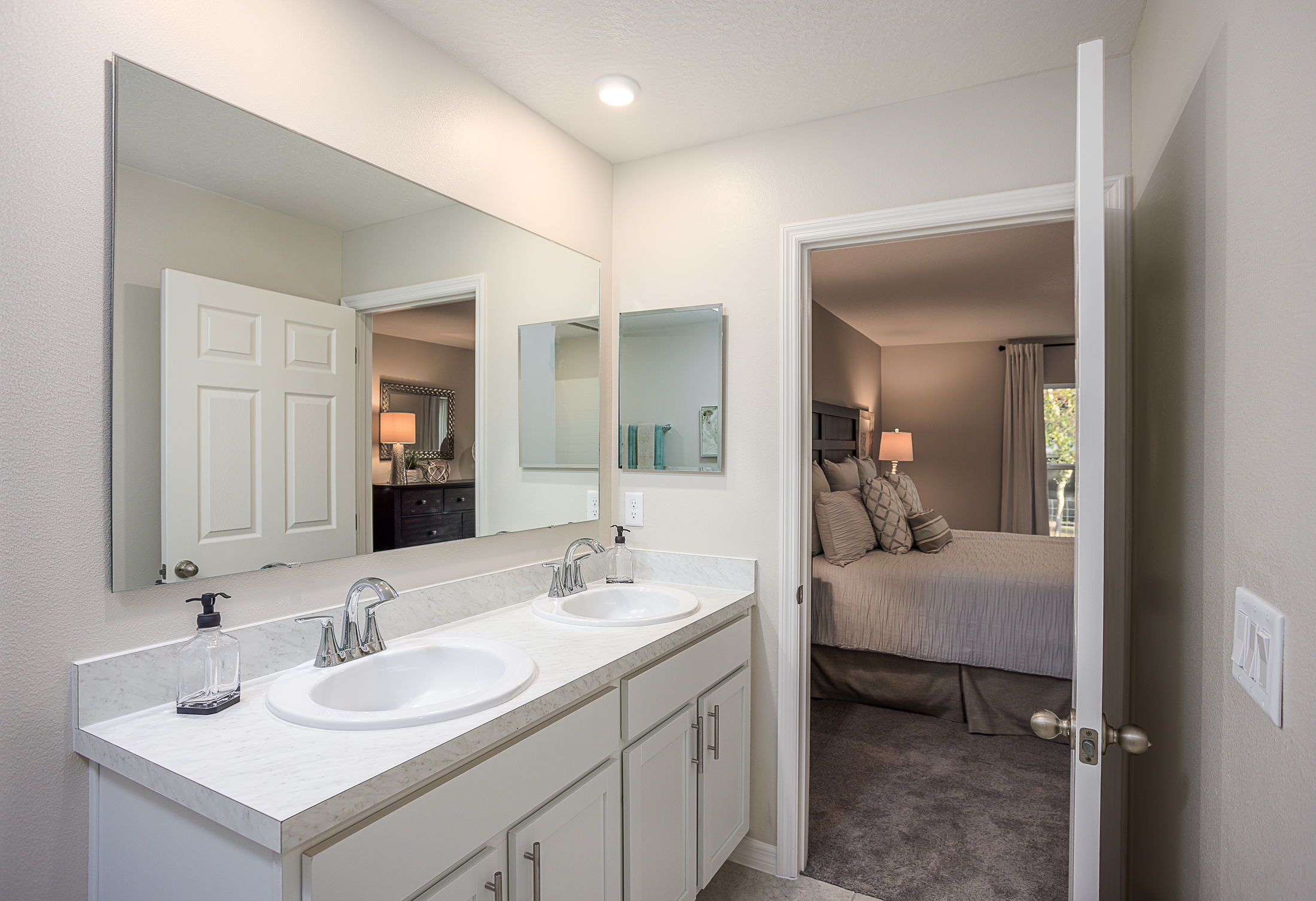 Modern bathroom with dual sinks and a large mirror adjacent to a stylish bedroom with ambient lighting.