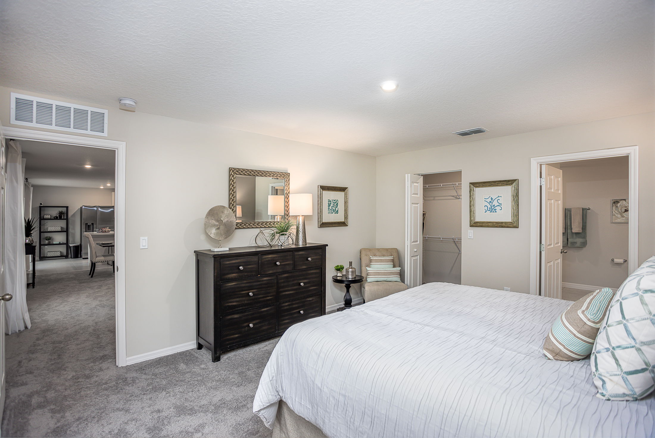 Modern bedroom with gray carpet, featuring a dark wooden dresser, abstract art, and a cozy chair, connected to a walk-in closet and en-suite bathroom.