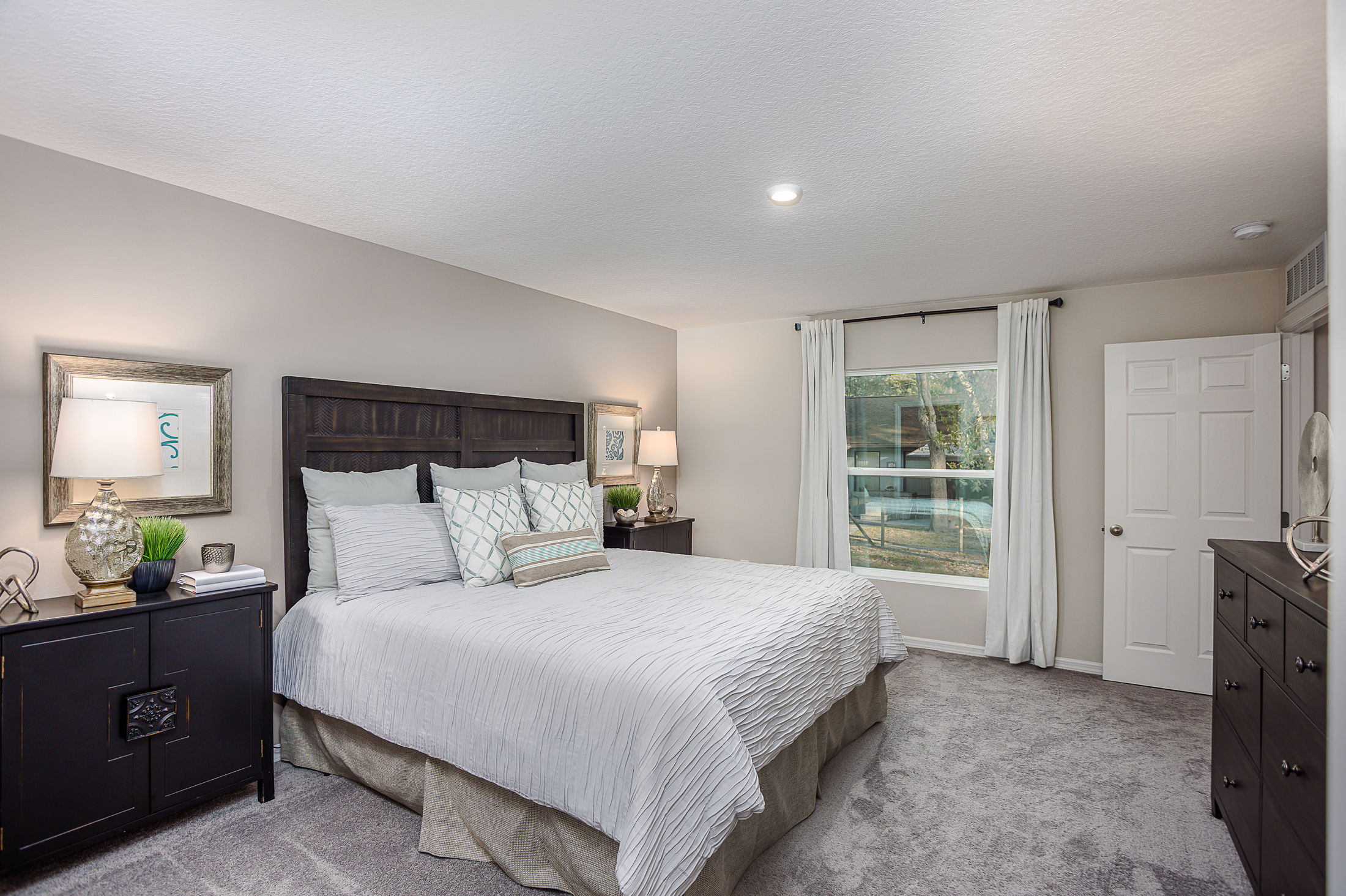 Elegant bedroom with a large bed, white bedding, dark wooden furniture, and a window with white curtains.