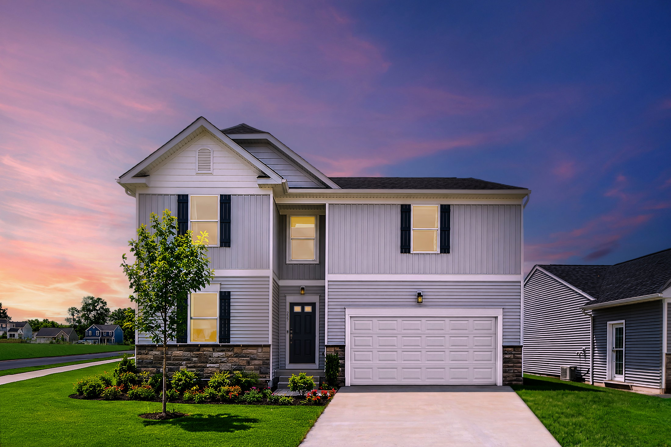 Two-story gray house with dark shutters, a landscaped front yard, and a double garage under a colorful sunset sky.