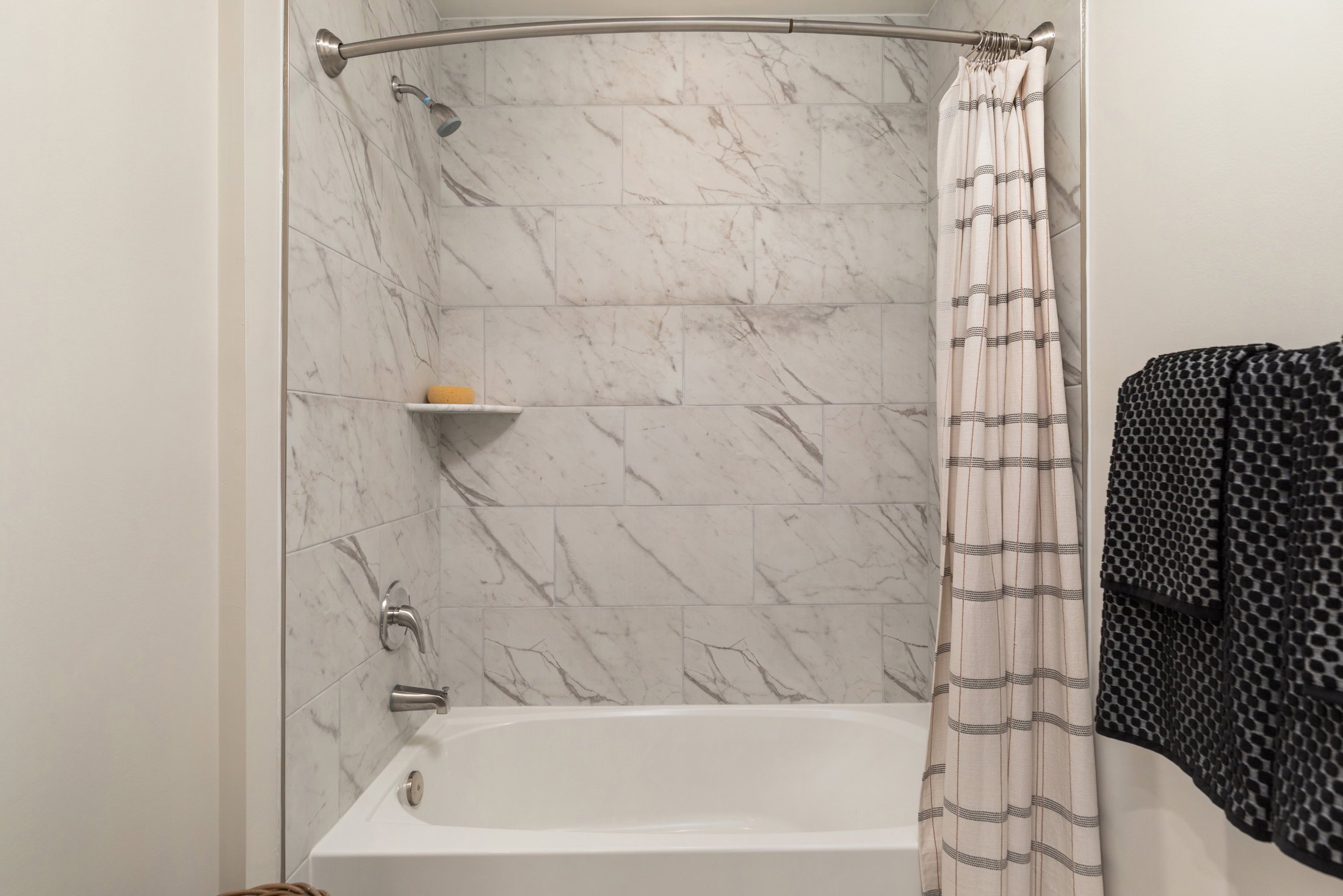 A modern bathroom featuring a marble-tiled shower area with a white bathtub, a shower curtain with a striped pattern, and a dark patterned towel hanging nearby.