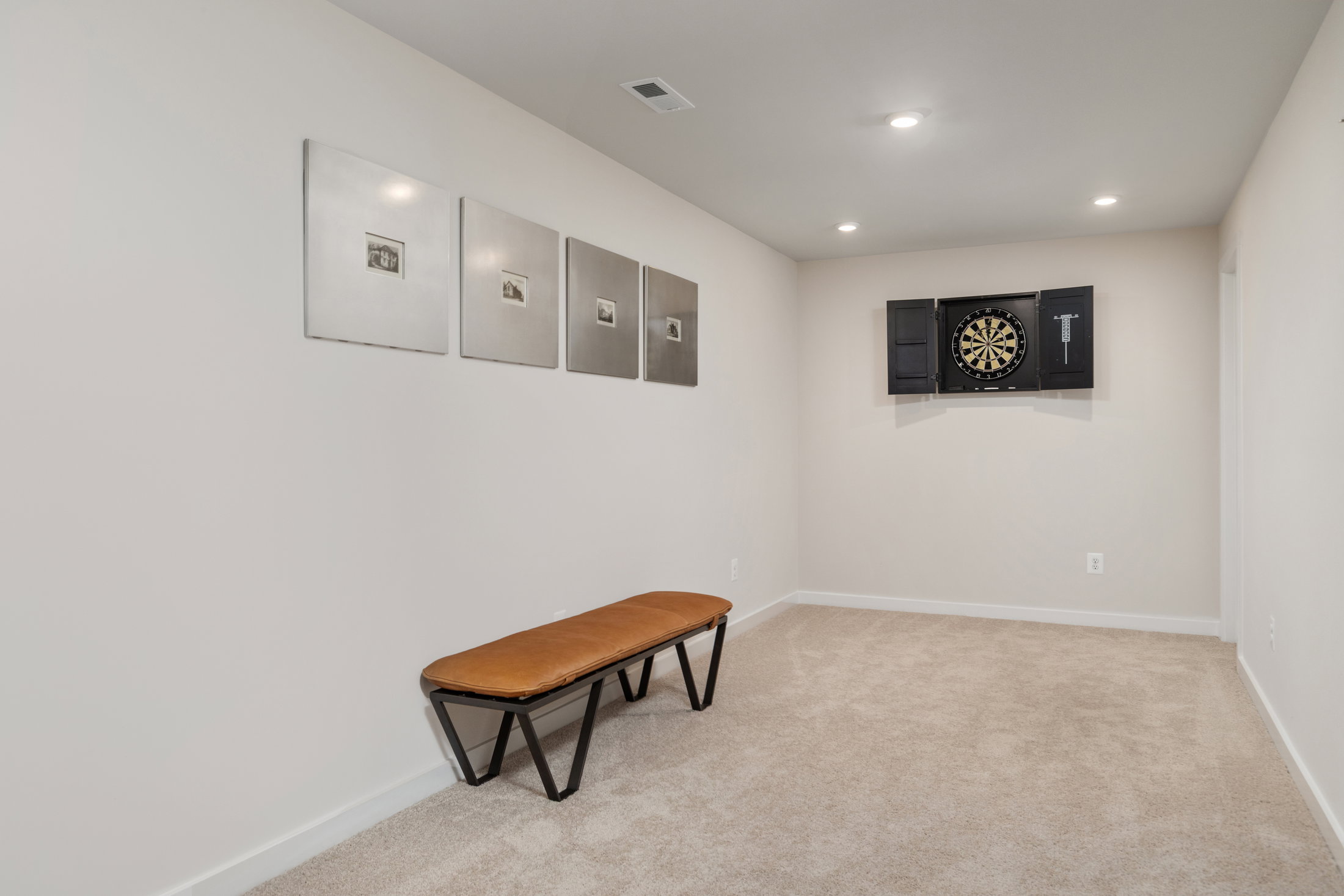 A room with beige walls, a dartboard on the wall, three framed artworks, and a brown bench.