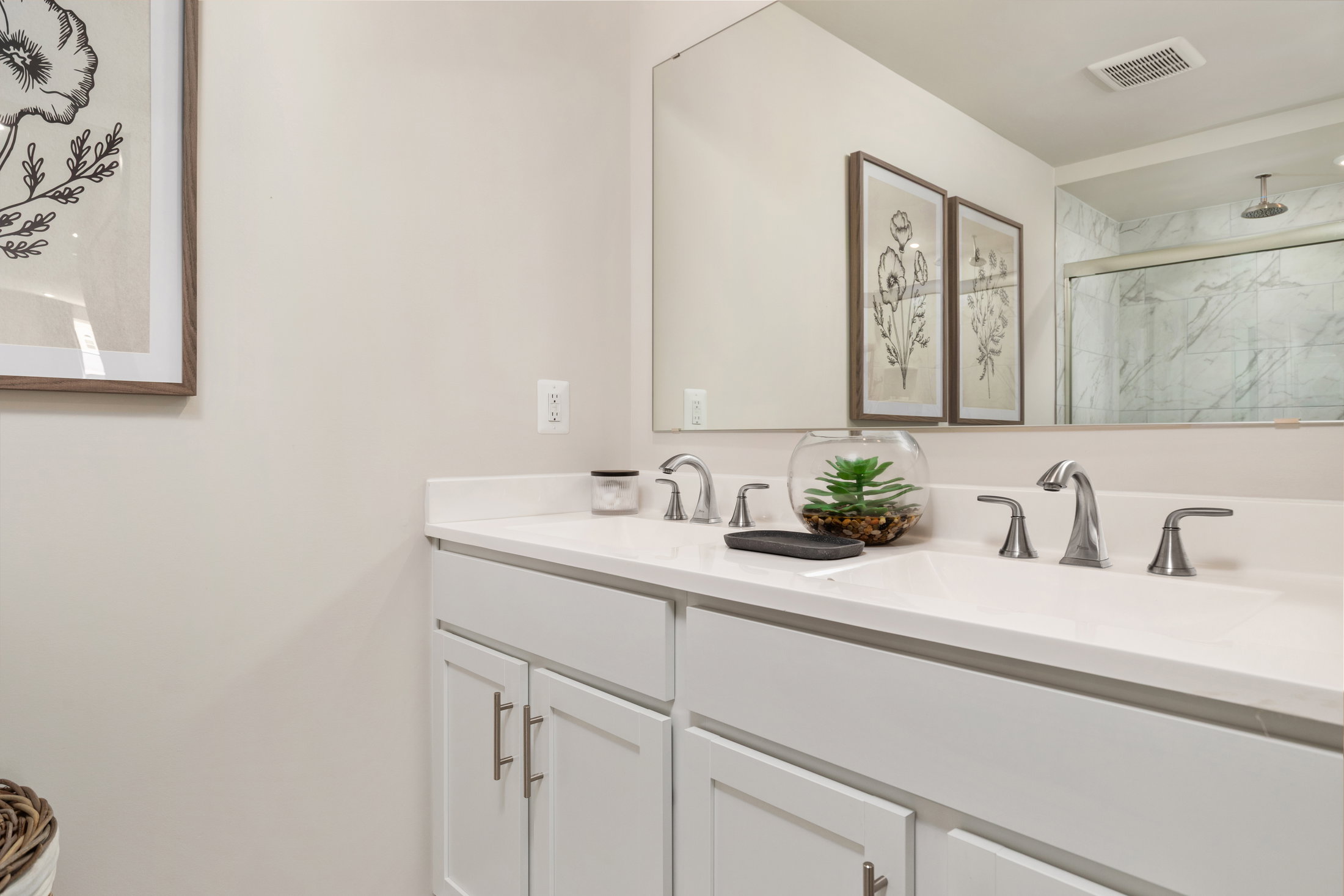 Vanity with a white countertop, two faucets, a decorative plant in a glass bowl, and framed wall art in a modern bathroom.