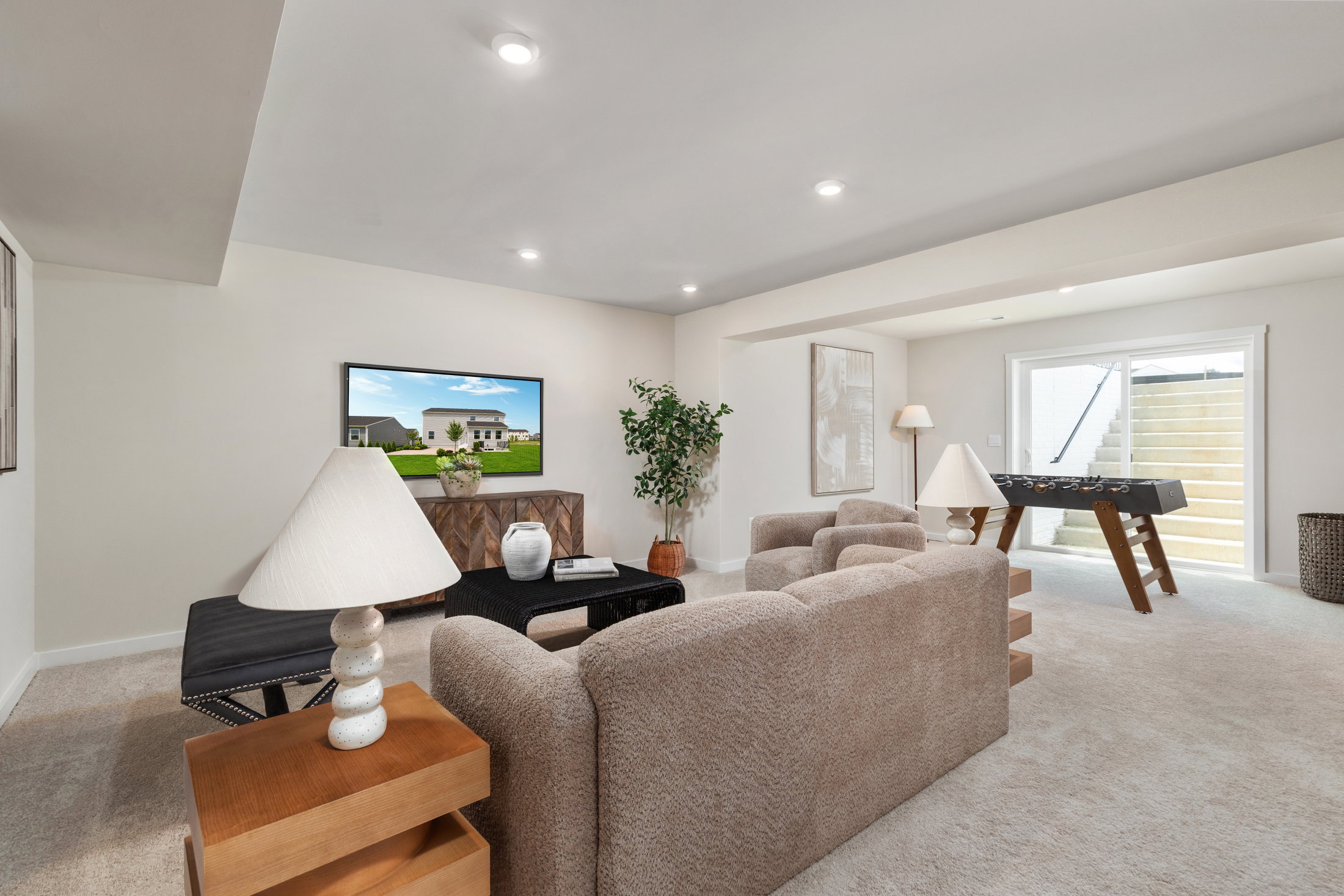 A light-filled basement living space with a beige couch, lamps, and a TV displaying a house image.
