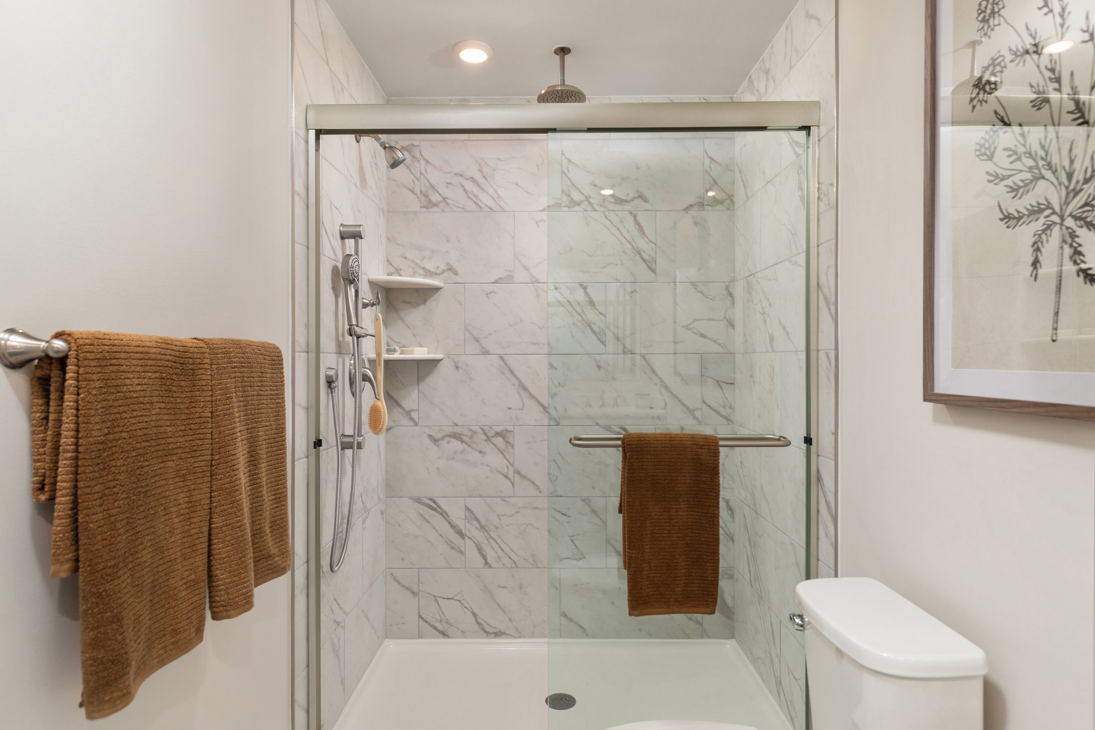 A modern shower with a glass enclosure, marble walls, and brown towels hanging on a rod.