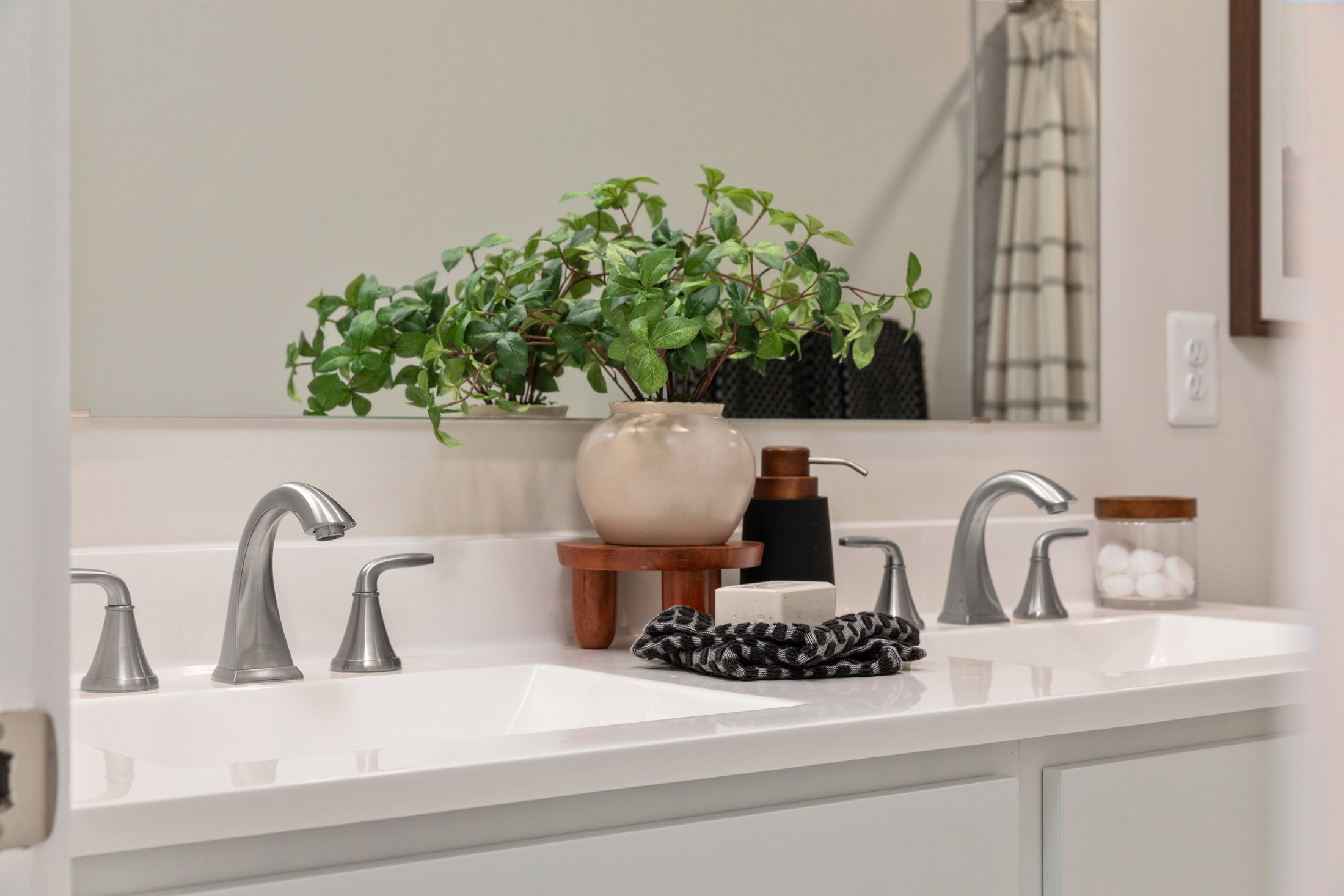 A clean bathroom sink with two faucets, a decorative plant in a pot, a soap dispenser, and a towel.