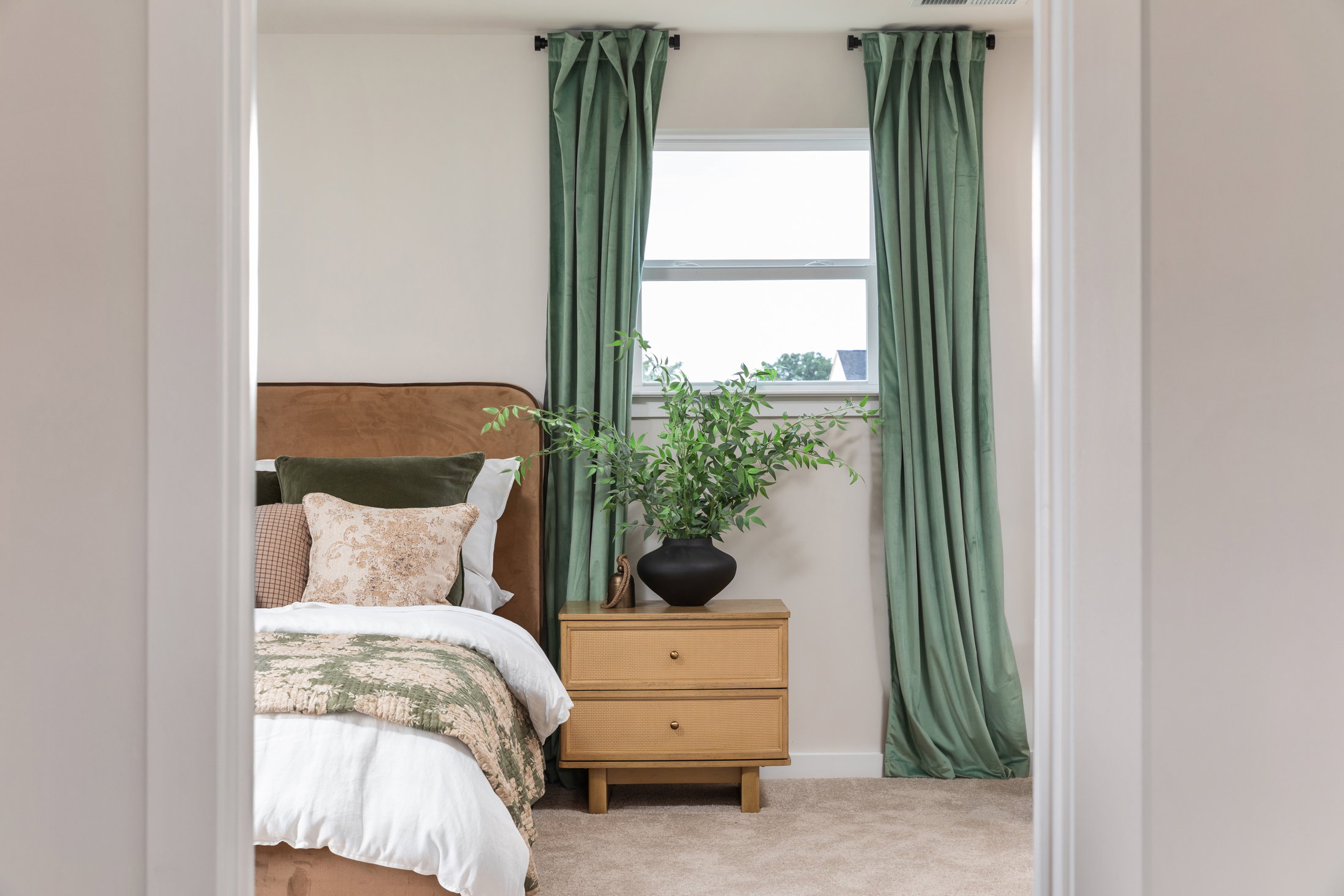 A cozy bedroom featuring a brown upholstered bed, decorative pillows, and a wooden nightstand with a black vase and green plant.