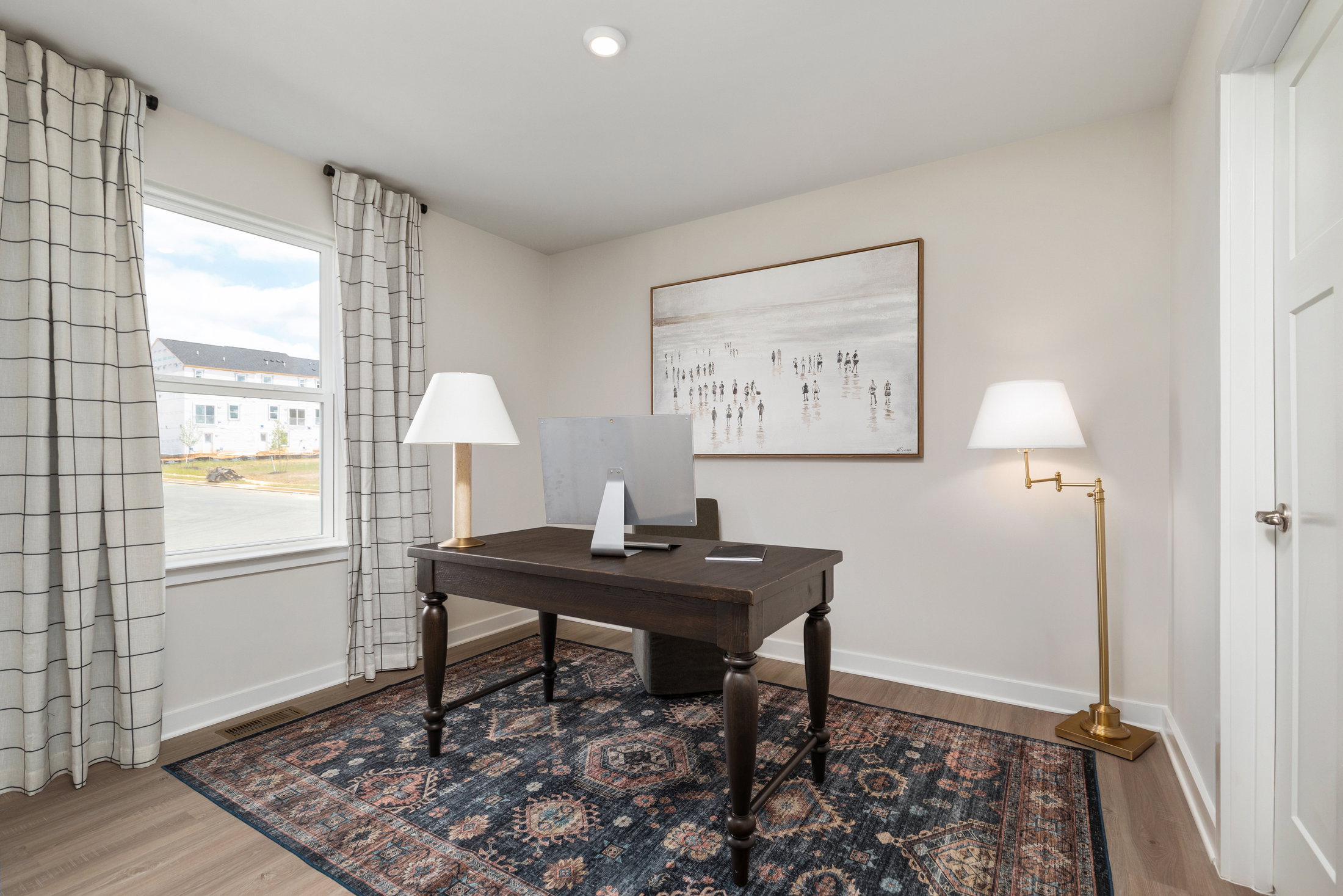 A modern home office featuring a wooden desk, a computer monitor, a gold floor lamp, and framed artwork, with large windows allowing natural light.