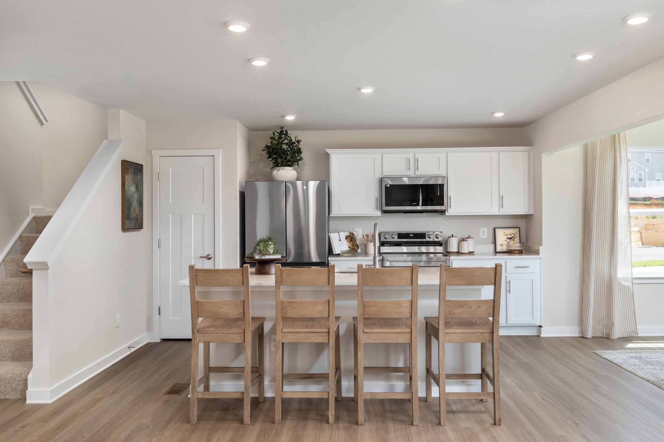 Modern kitchen with a large island, six wooden stools, stainless steel appliances, and light-colored cabinetry.