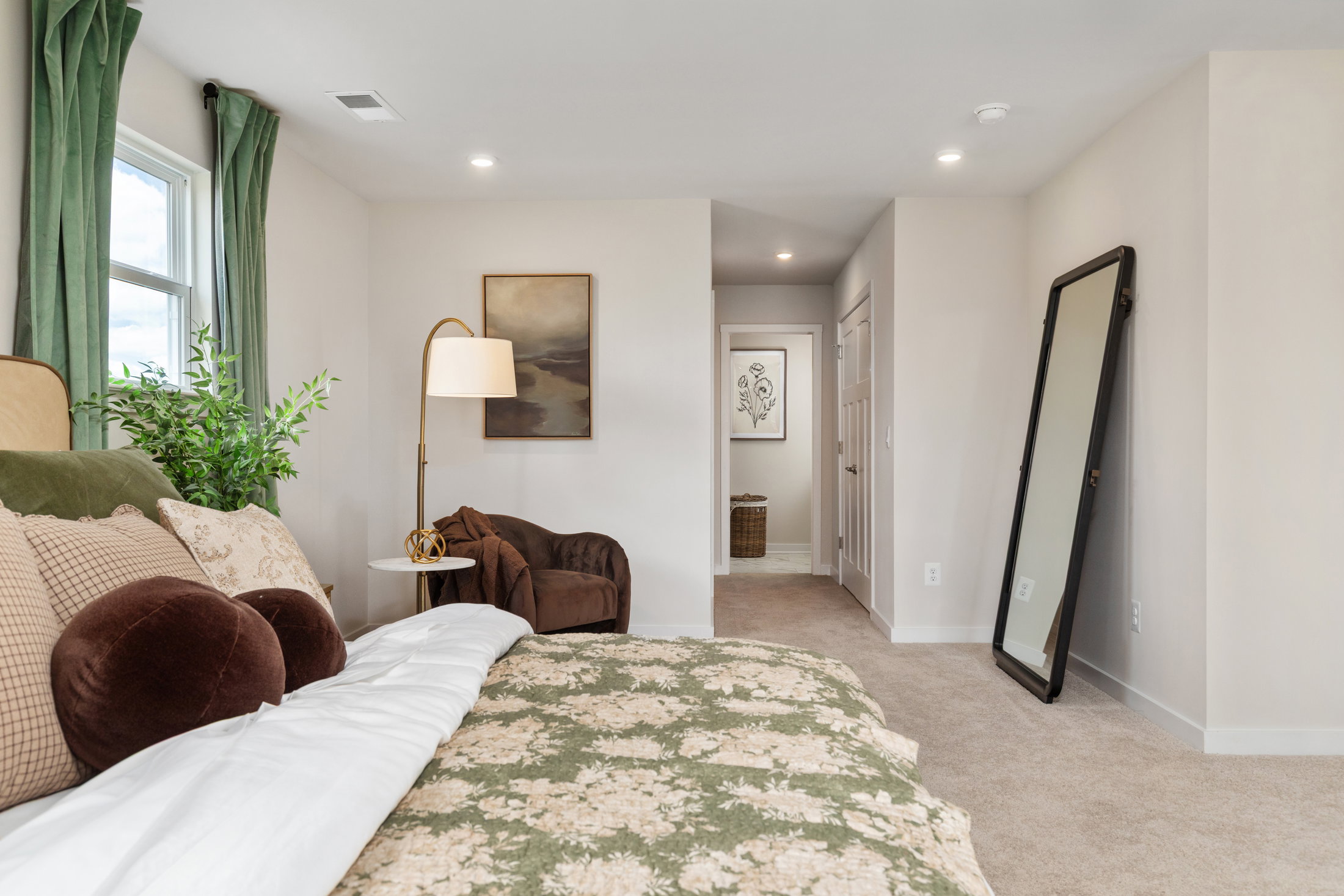 A cozy bedroom featuring a bed with a floral blanket, a floor lamp, a brown armchair, and a large mirror.