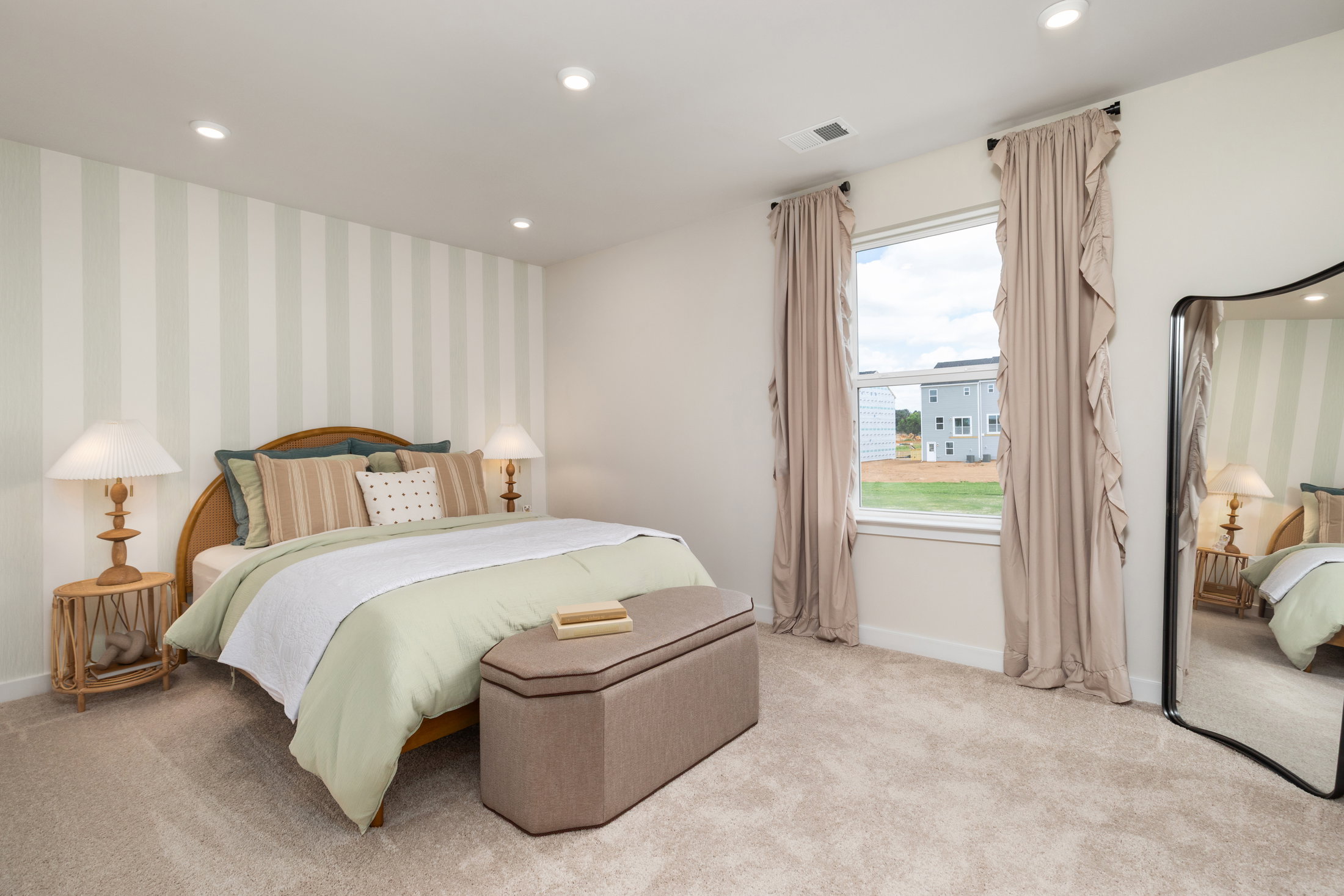 A cozy bedroom featuring a bed with pastel green bedding, a decorative bench, two lamps, and large windows with curtains.