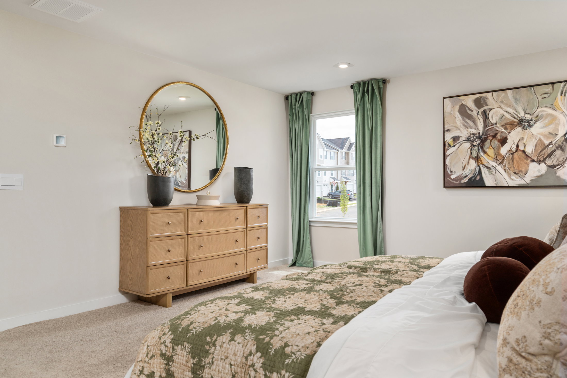 A cozy bedroom featuring a wooden dresser with decorative items, a round mirror, floral artwork, and green curtains framing a window.