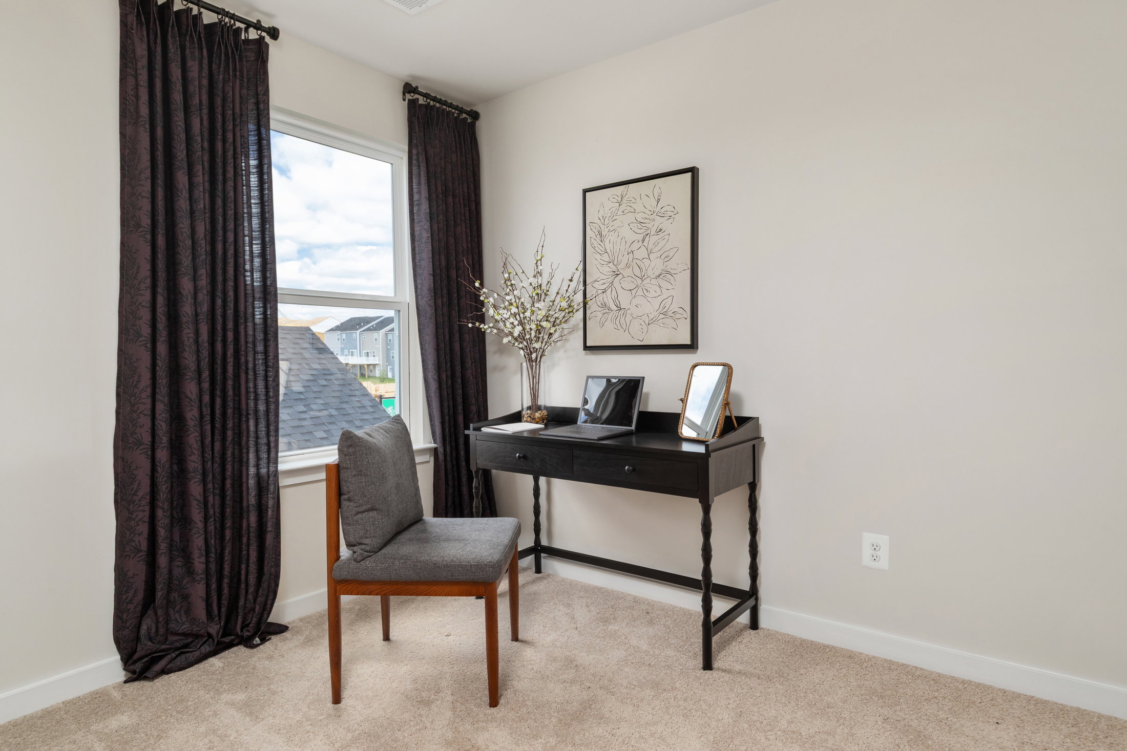 A small desk with a chair, a framed artwork on the wall, and a vase with decorative plants in a well-lit room.