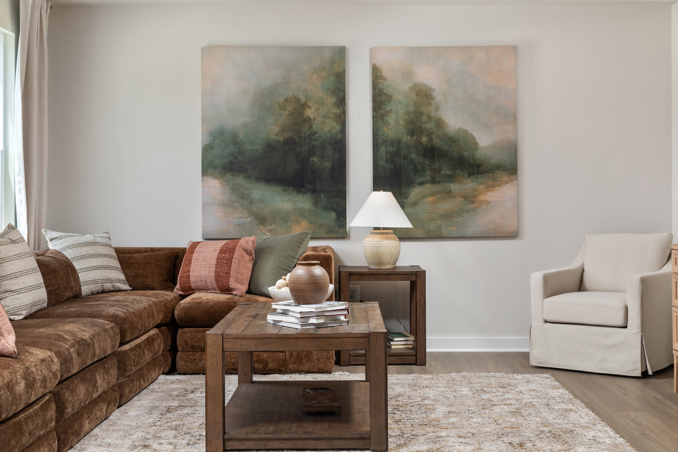 A cozy living room featuring a brown sectional sofa, a wooden coffee table with books, a ceramic vase, and wall art depicting a serene landscape.
