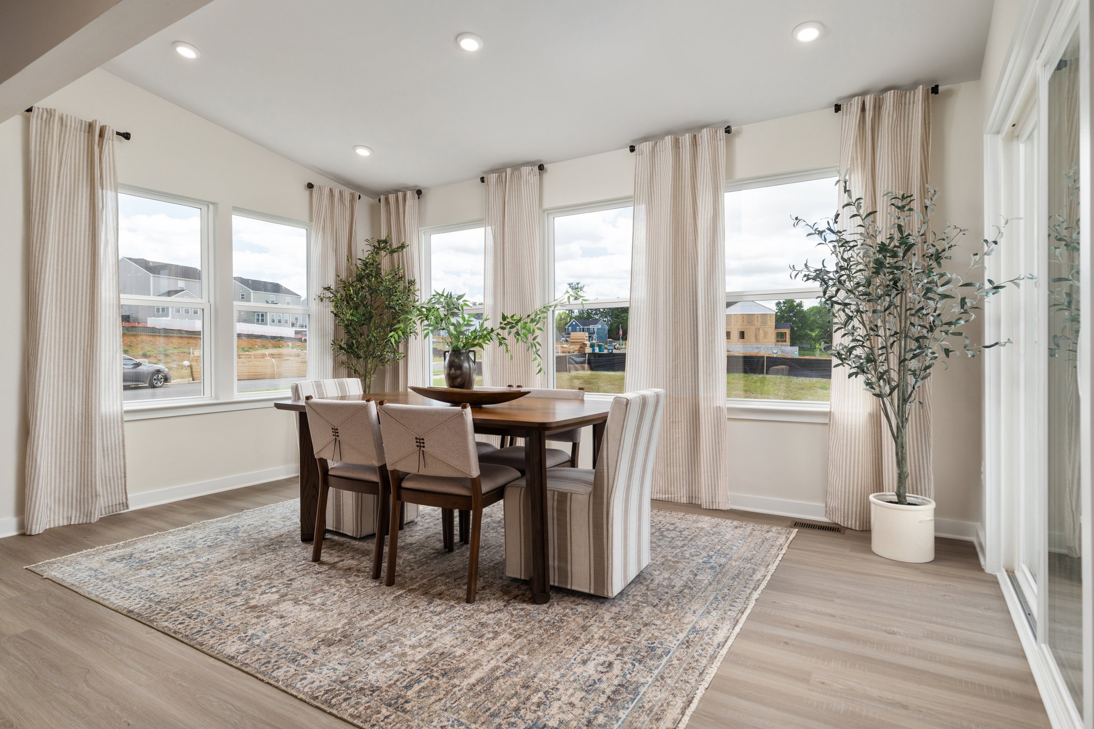 Dining area with a wooden table surrounded by beige upholstered chairs, large windows with light curtains, and potted plants.