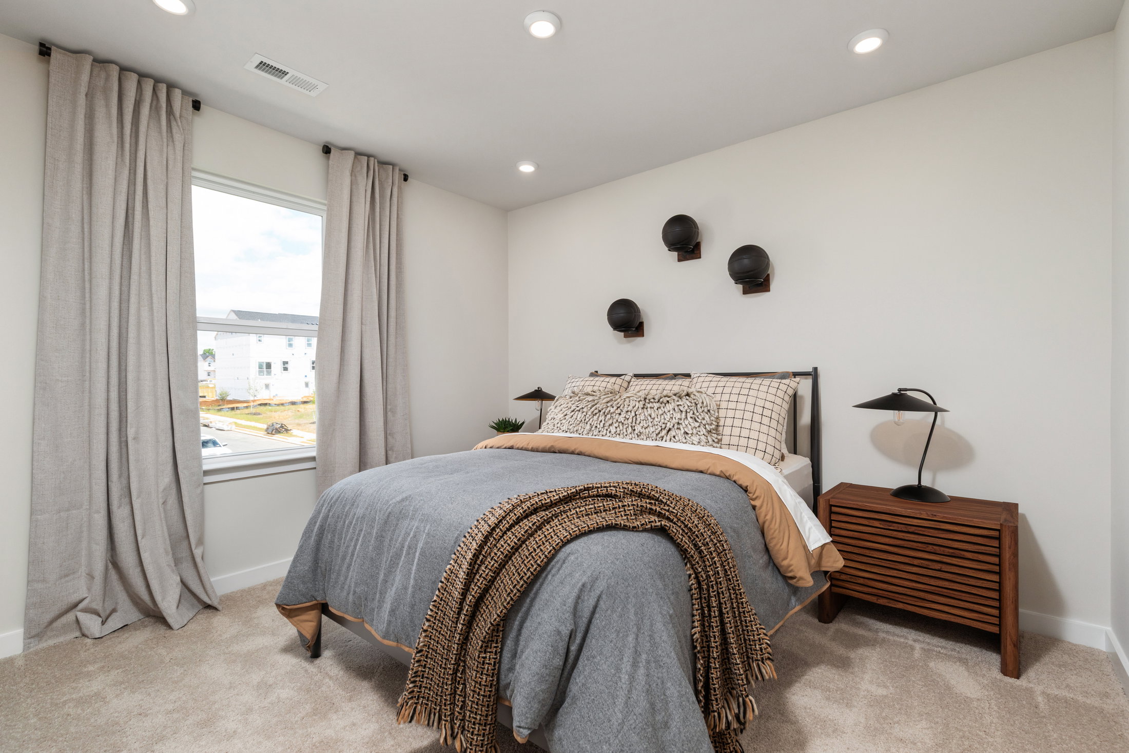 A well-decorated bedroom featuring a bed with grey and beige bedding, a wooden nightstand, and wall sconces, with large windows allowing natural light.