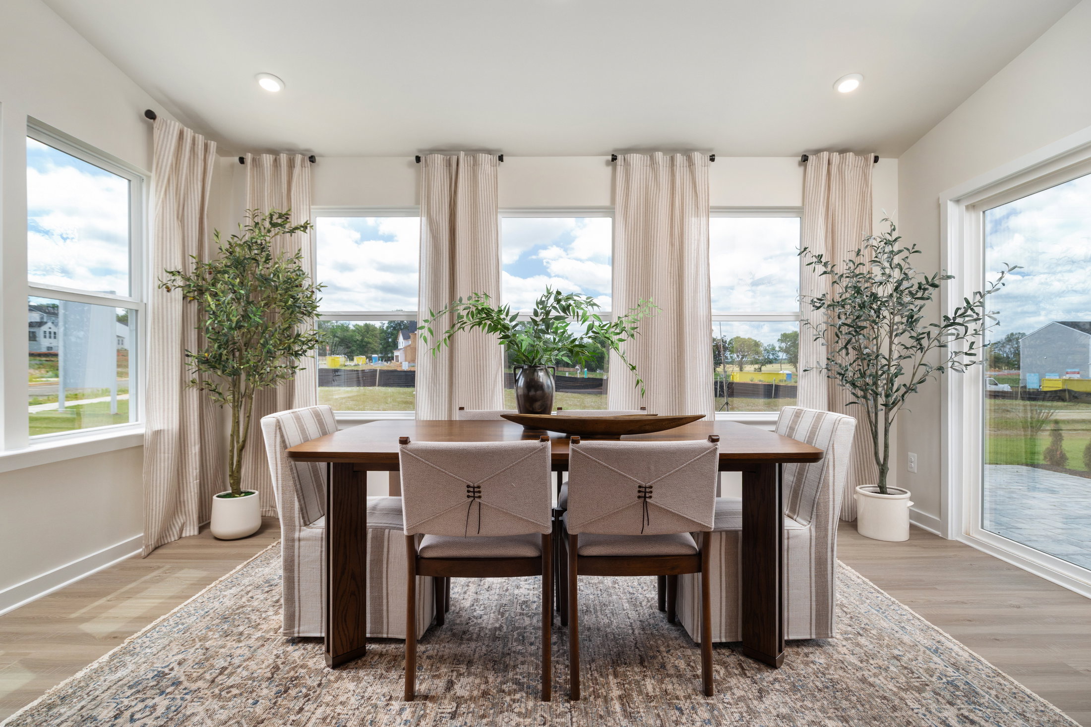 Bright dining area featuring a wooden table, four upholstered chairs, and large windows with light curtains overlooking greenery.