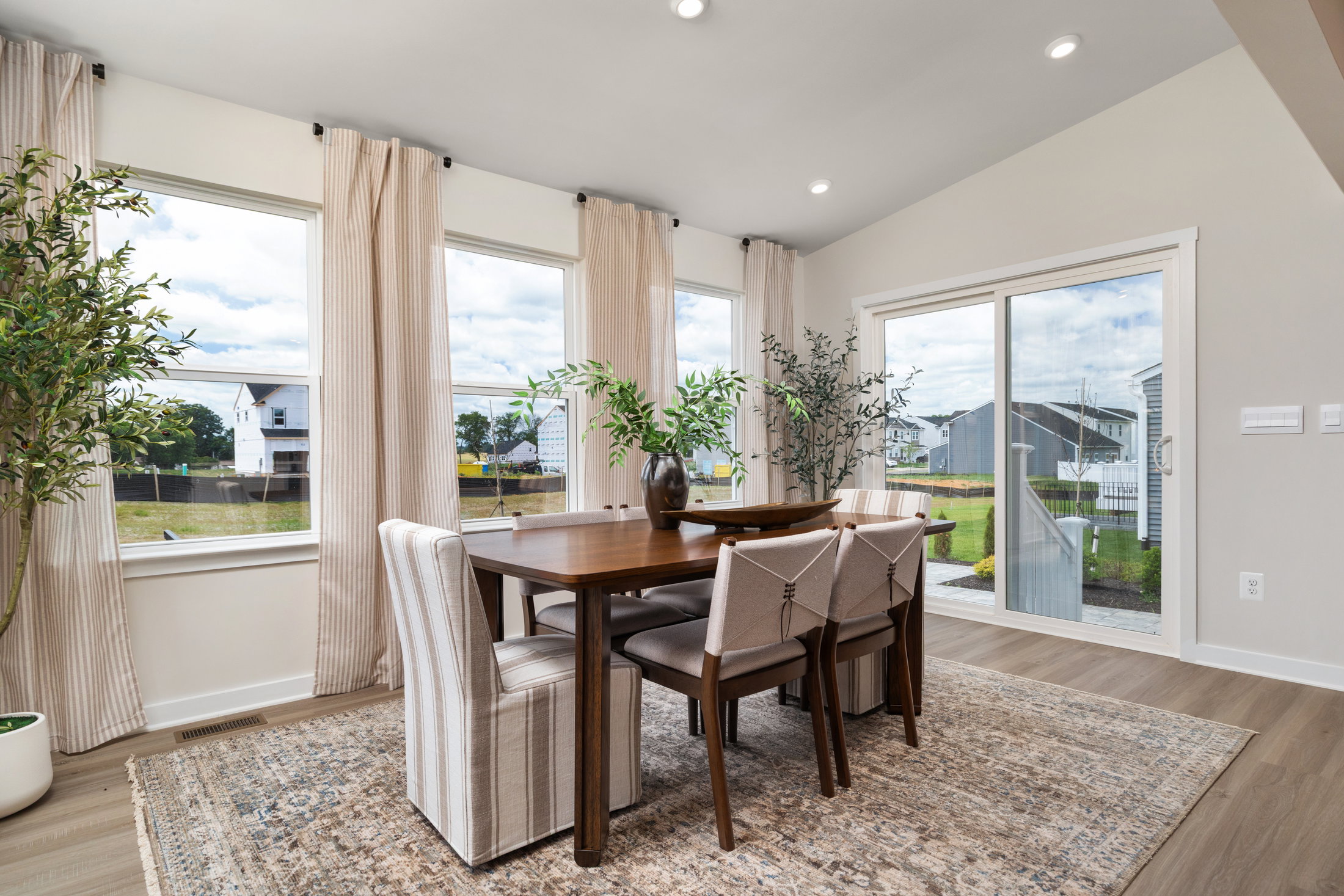 A dining area featuring a wooden table surrounded by upholstered chairs, large windows with sheer curtains, and potted plants.