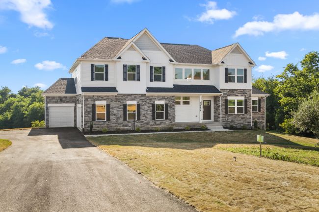 Two-story suburban house with a stone facade and attached garage, surrounded by a freshly mowed lawn and trees under a clear blue sky.