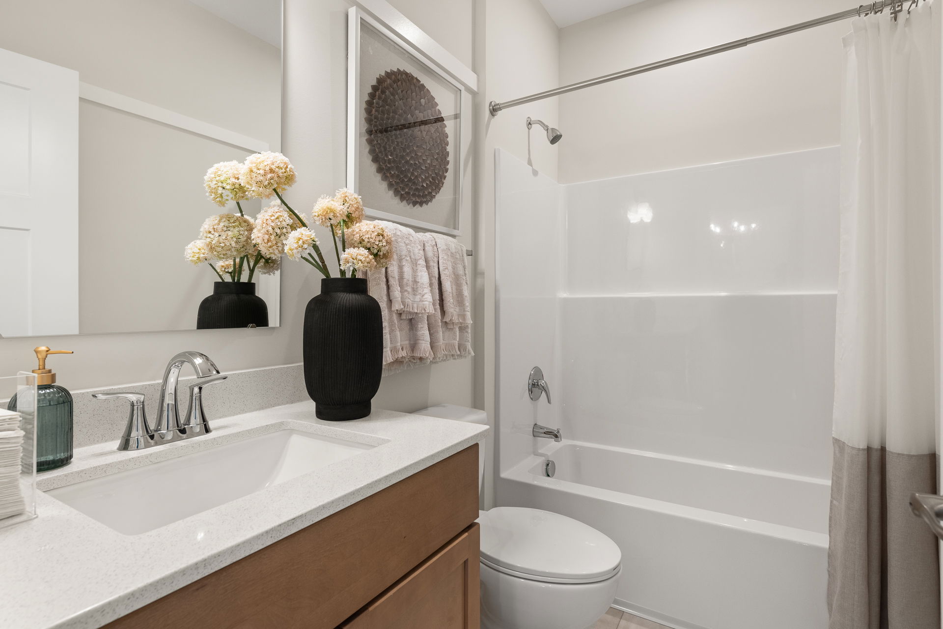 Modern bathroom with white countertops, a sleek sink, black vase with flowers, and a glass shower-tub combo.