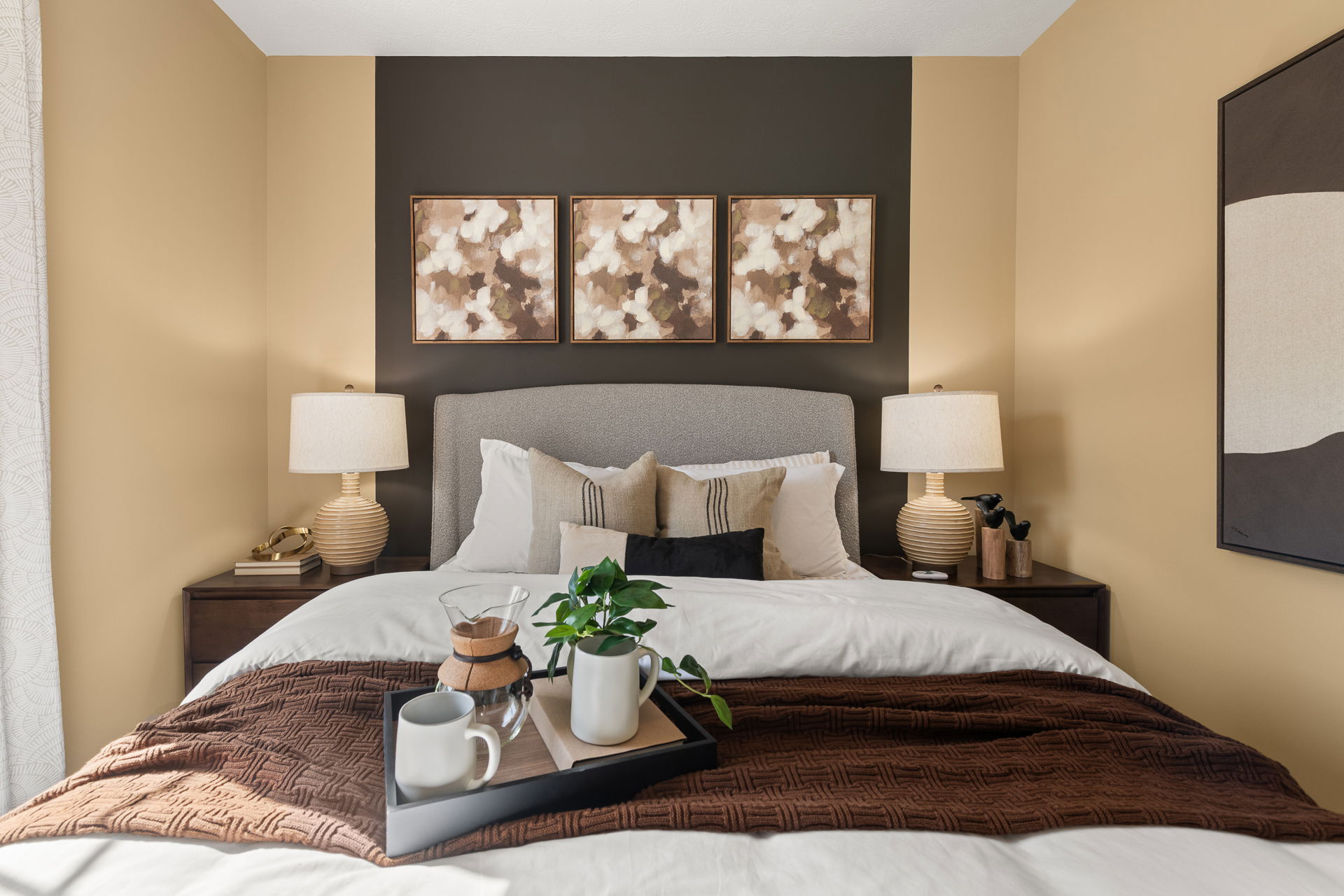 Stylish modern bedroom with a comfortable bed, decorative pillows, and a tray featuring coffee and a plant, accented by wall art and warm lighting.