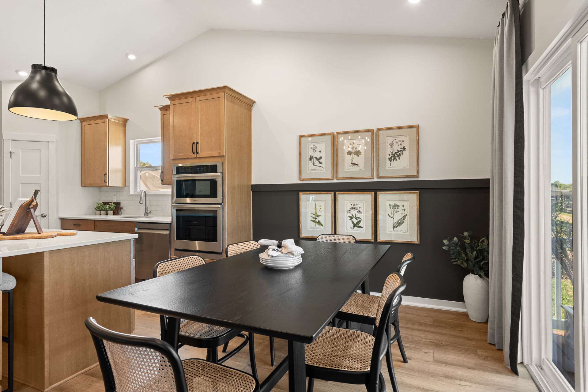 Modern kitchen and dining area featuring a black dining table with woven chairs, wooden cabinetry, built-in appliances, and botanical artwork on the wall.