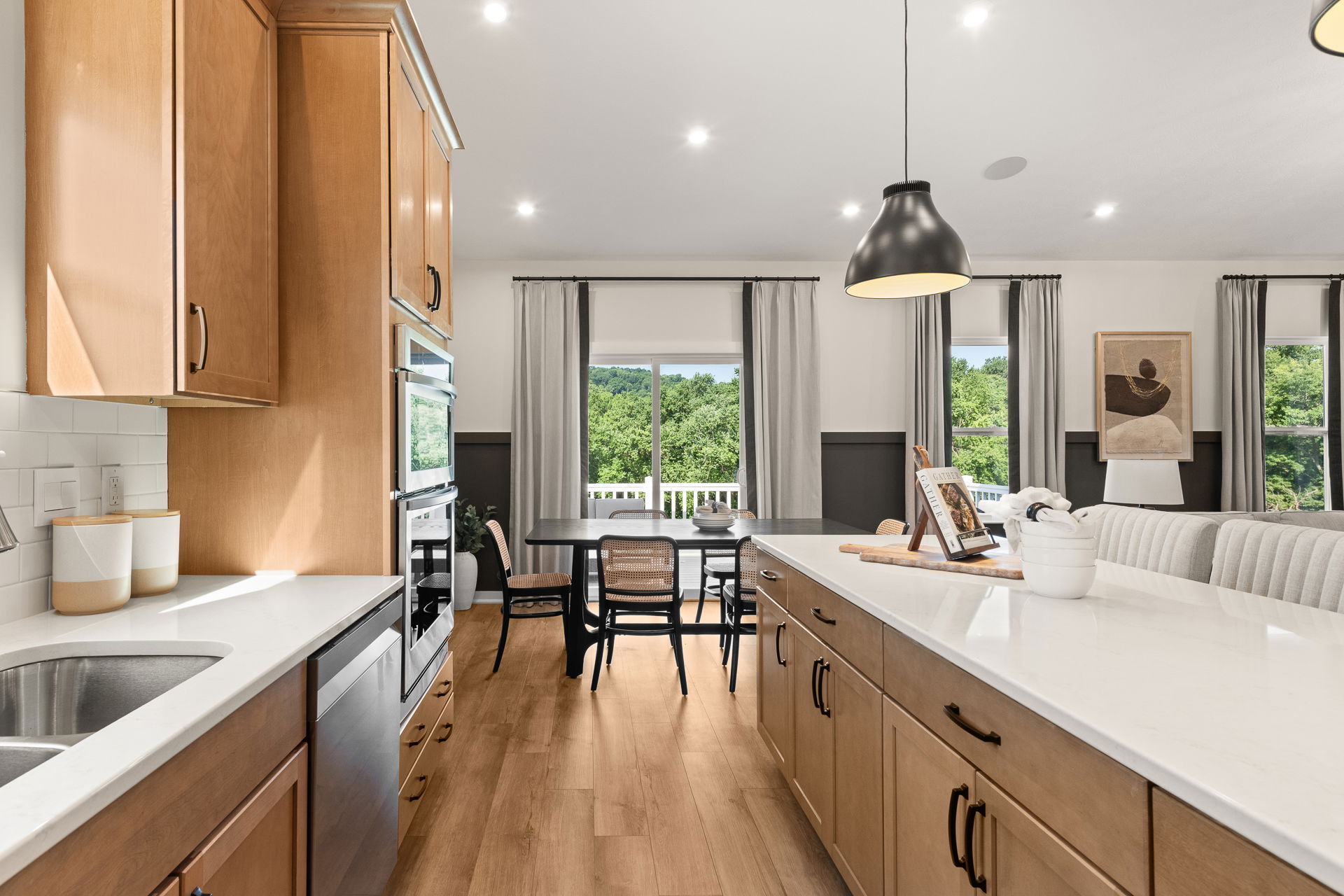Modern kitchen interior with light wood cabinets, white countertops, and a dining area overlooking a scenic view through large windows.