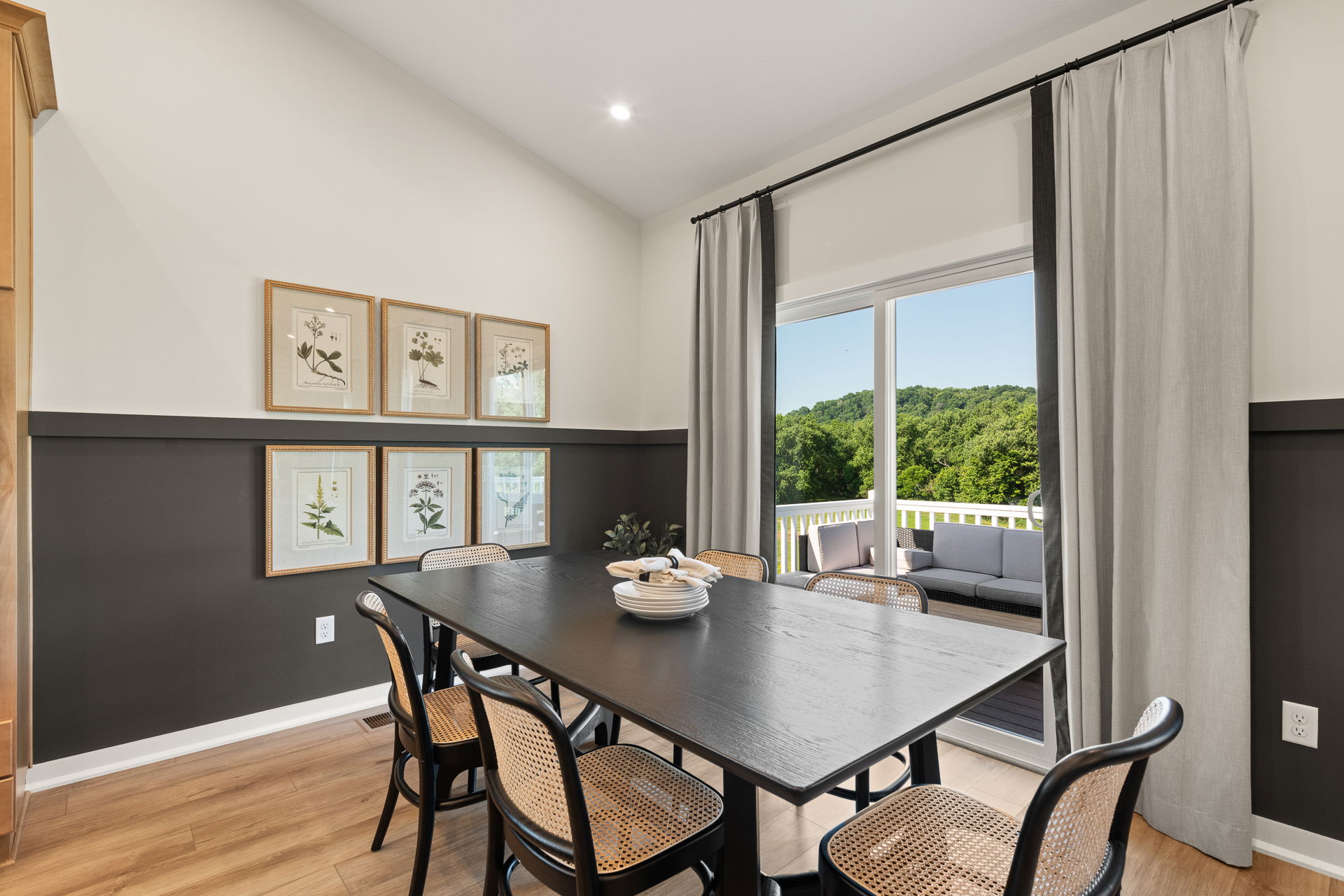 Elegant dining room with black table, wicker chairs, botanical wall art, and sliding glass doors leading to a scenic patio view.