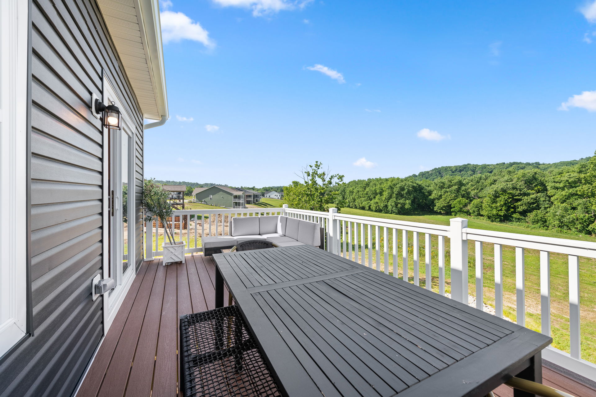 Modern outdoor deck with gray siding, railing, and patio furniture, overlooking a scenic landscape and clear blue sky.