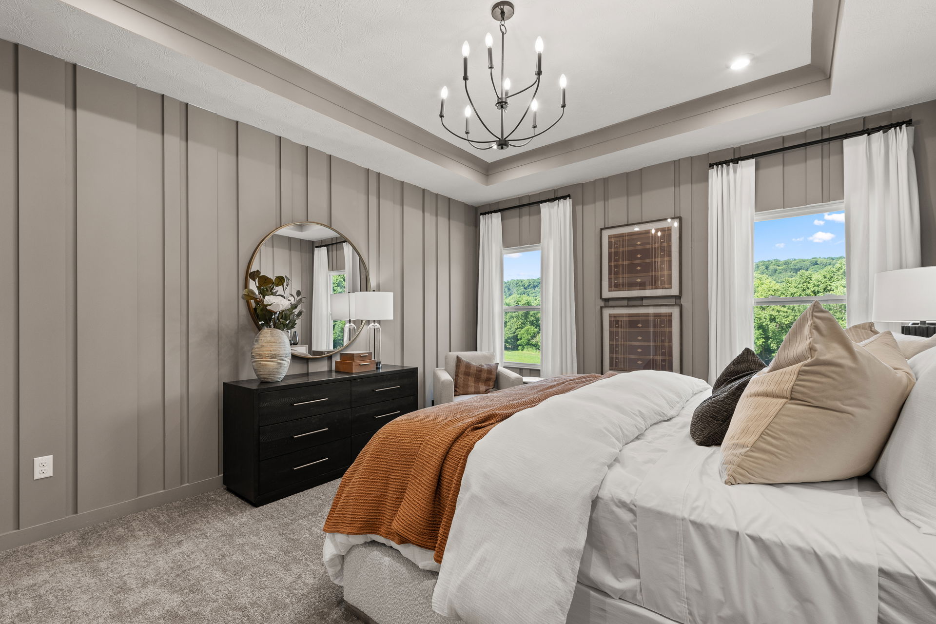 Modern bedroom with elegant chandelier, neutral tones, and large windows overlooking lush greenery.