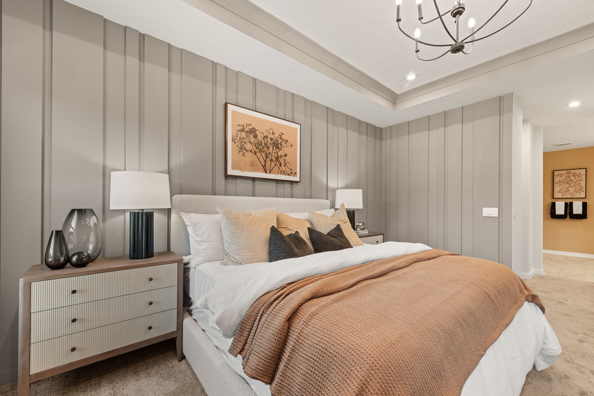 Modern bedroom with a cozy bed featuring neutral pillows and an orange blanket, flanked by stylish nightstands and soft lighting, under a chic chandelier.