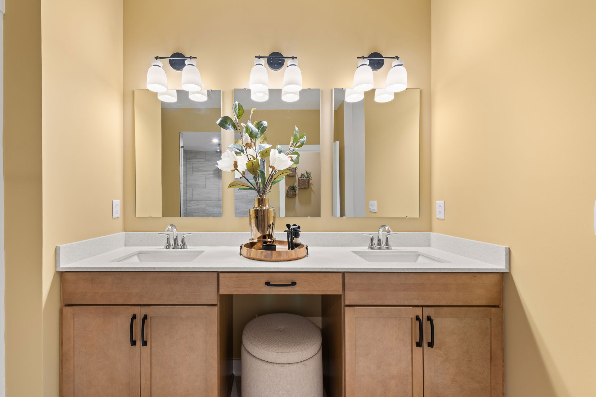 Modern bathroom vanity with dual sinks, light wood cabinets, large mirrors, elegant lighting, and decorative vase with flowers.