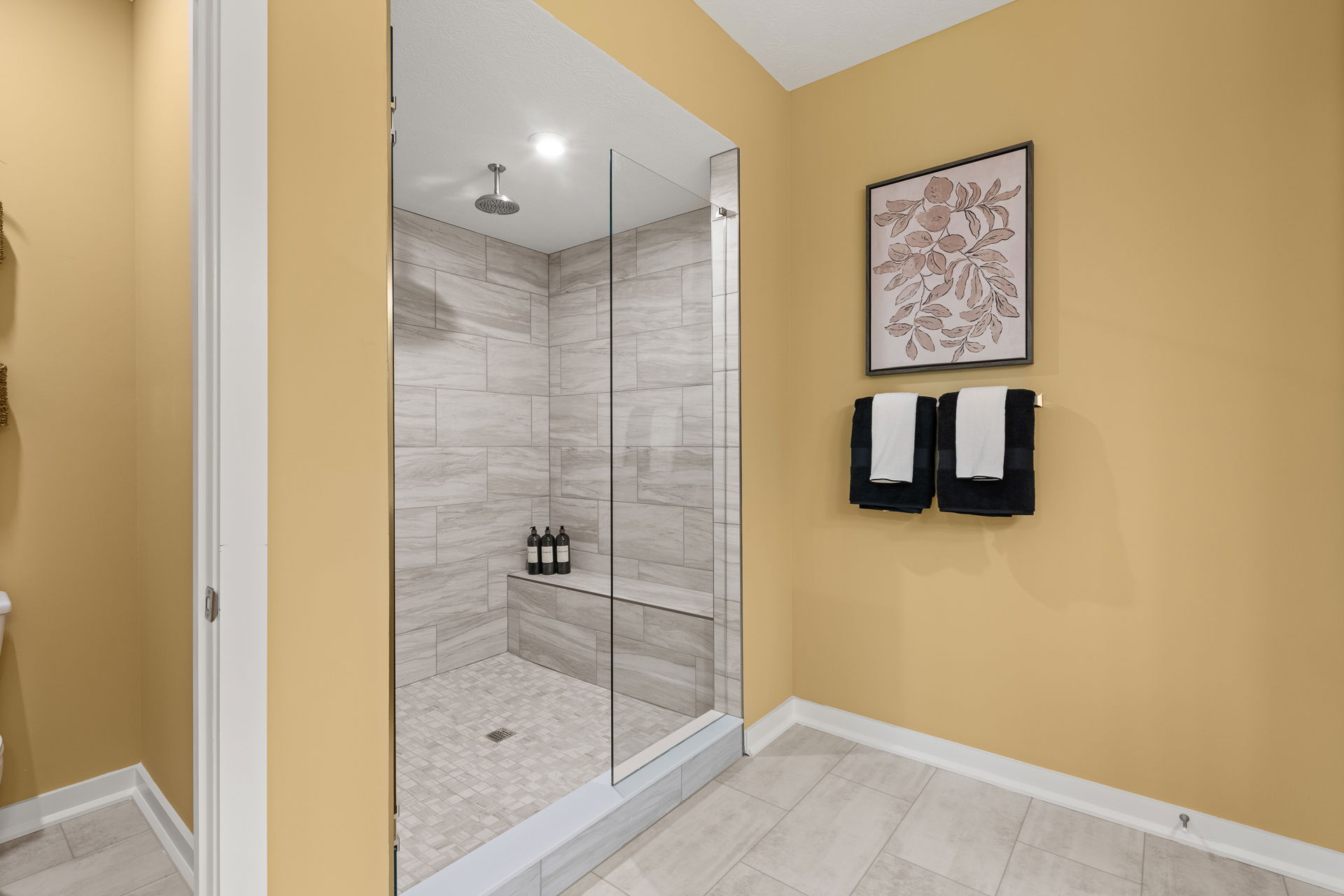 Modern bathroom with a glass-enclosed shower, beige tile walls, and folded towels on a yellow-painted wall.