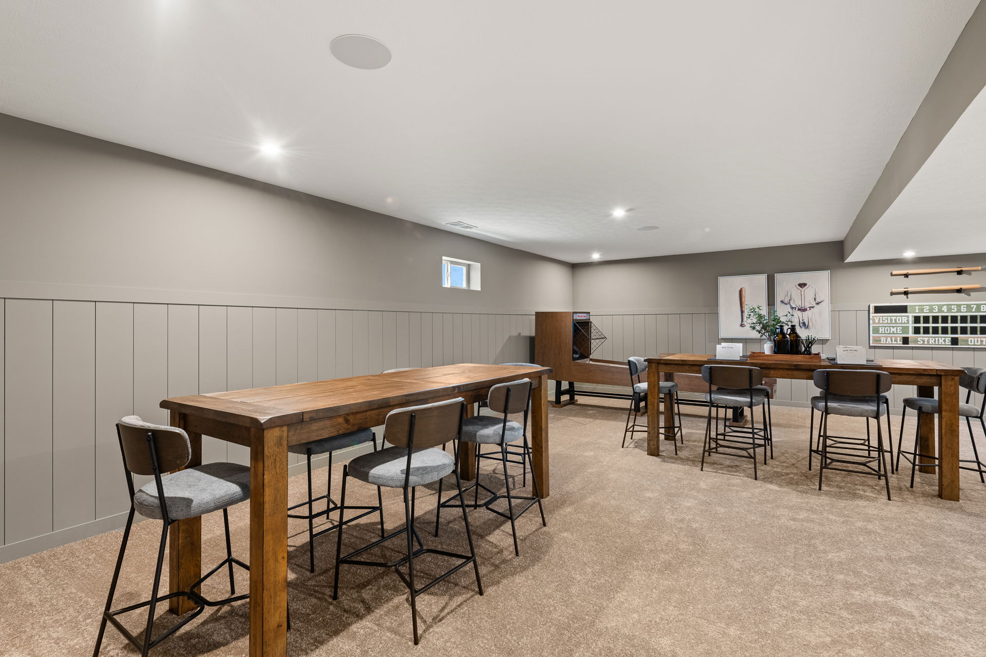 Modern basement game room with wooden high tables, gray chairs, and baseball-themed decor.