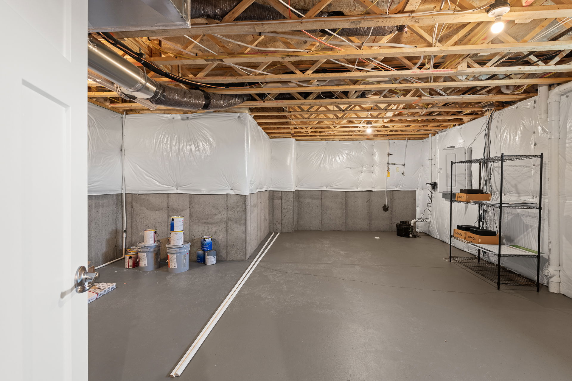 Unfinished basement with exposed wooden beams, white plastic insulation, paint cans, and a metal storage shelf.