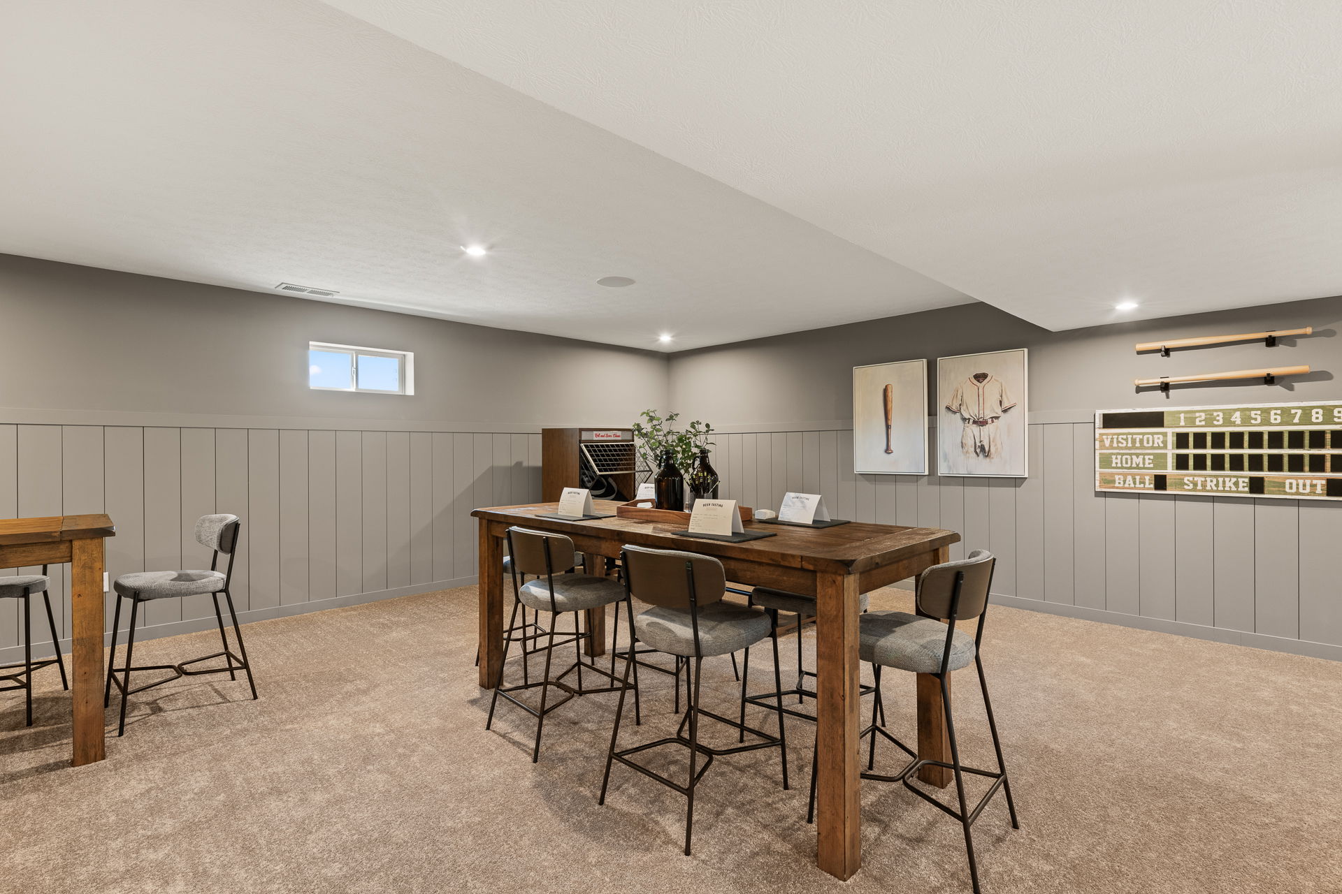 Modern basement game room with wooden table, bar stools, baseball-themed decor, and scoreboard on gray paneled walls.