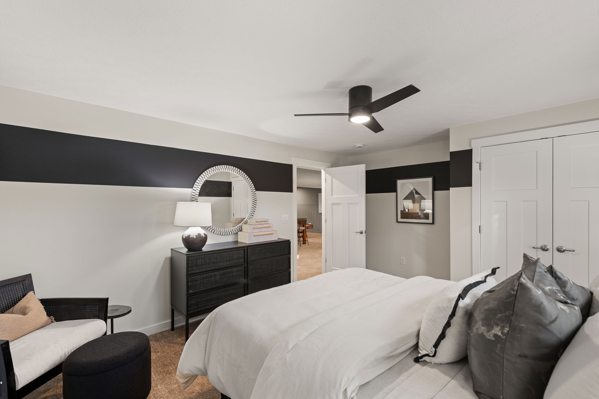 Modern bedroom with sleek black furniture, white bedding, decorative mirror, and a ceiling fan.